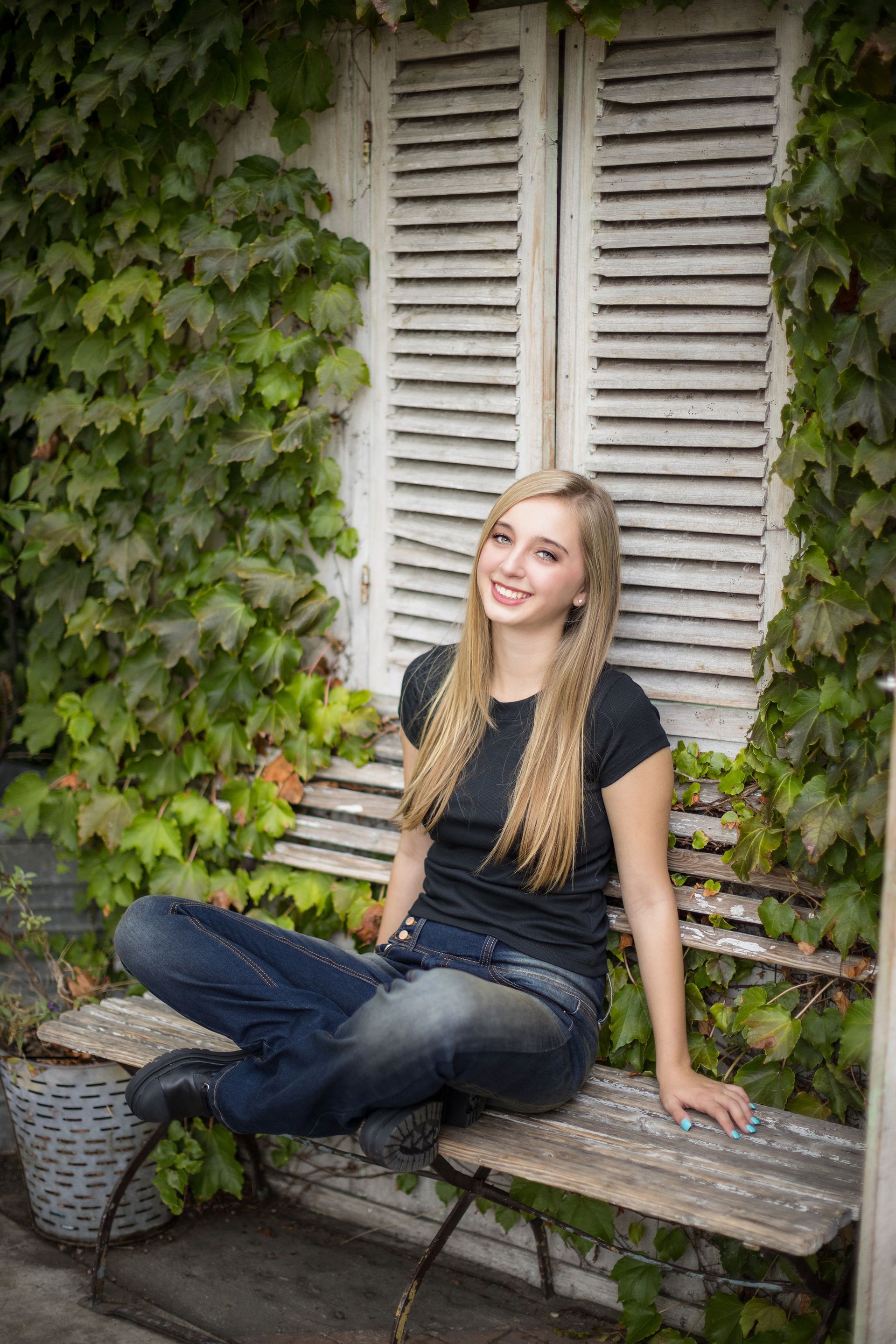 A young woman is sitting on a wooden bench in front of a window with shutters.