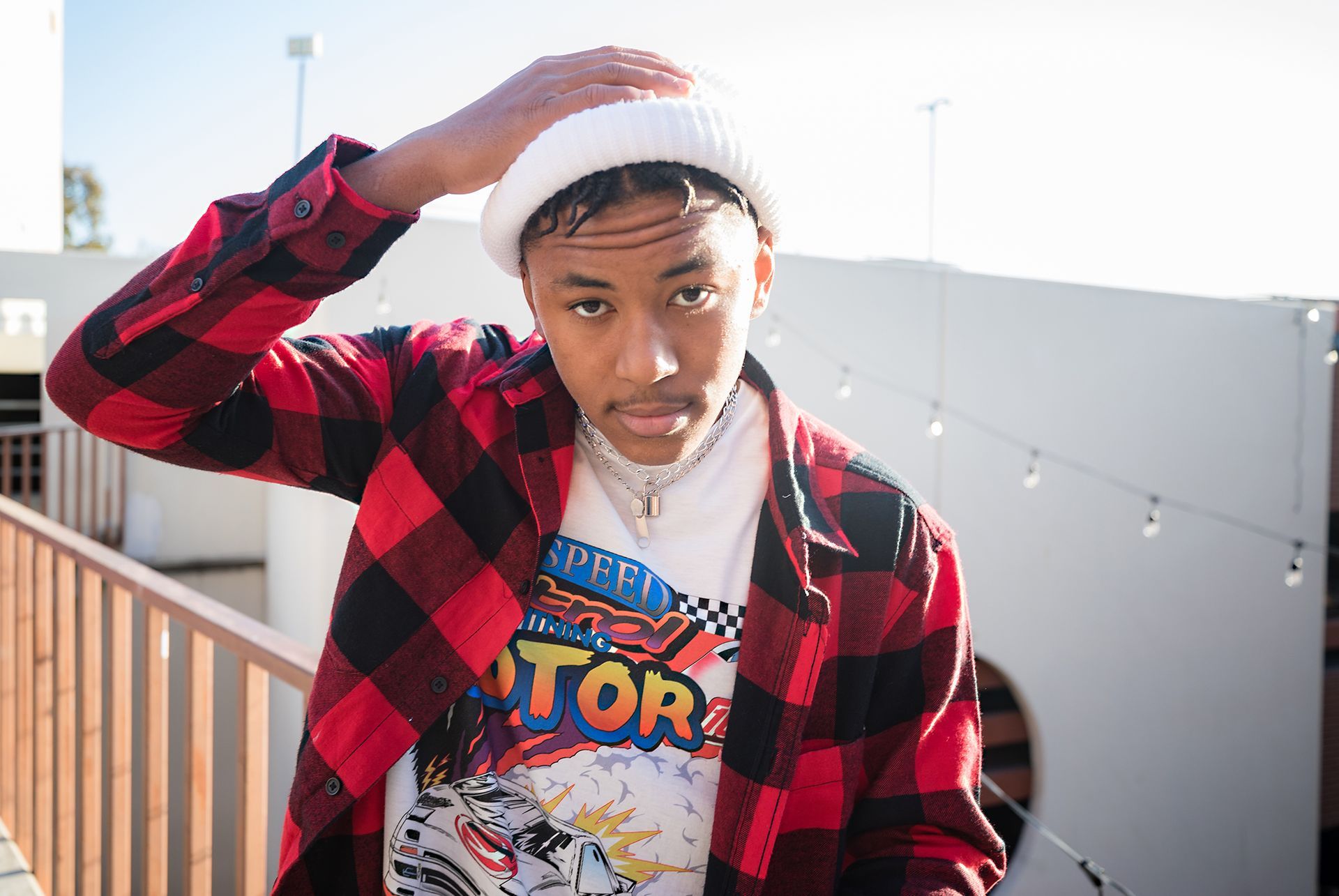 A young man wearing a plaid shirt and a white hat is standing on a balcony.