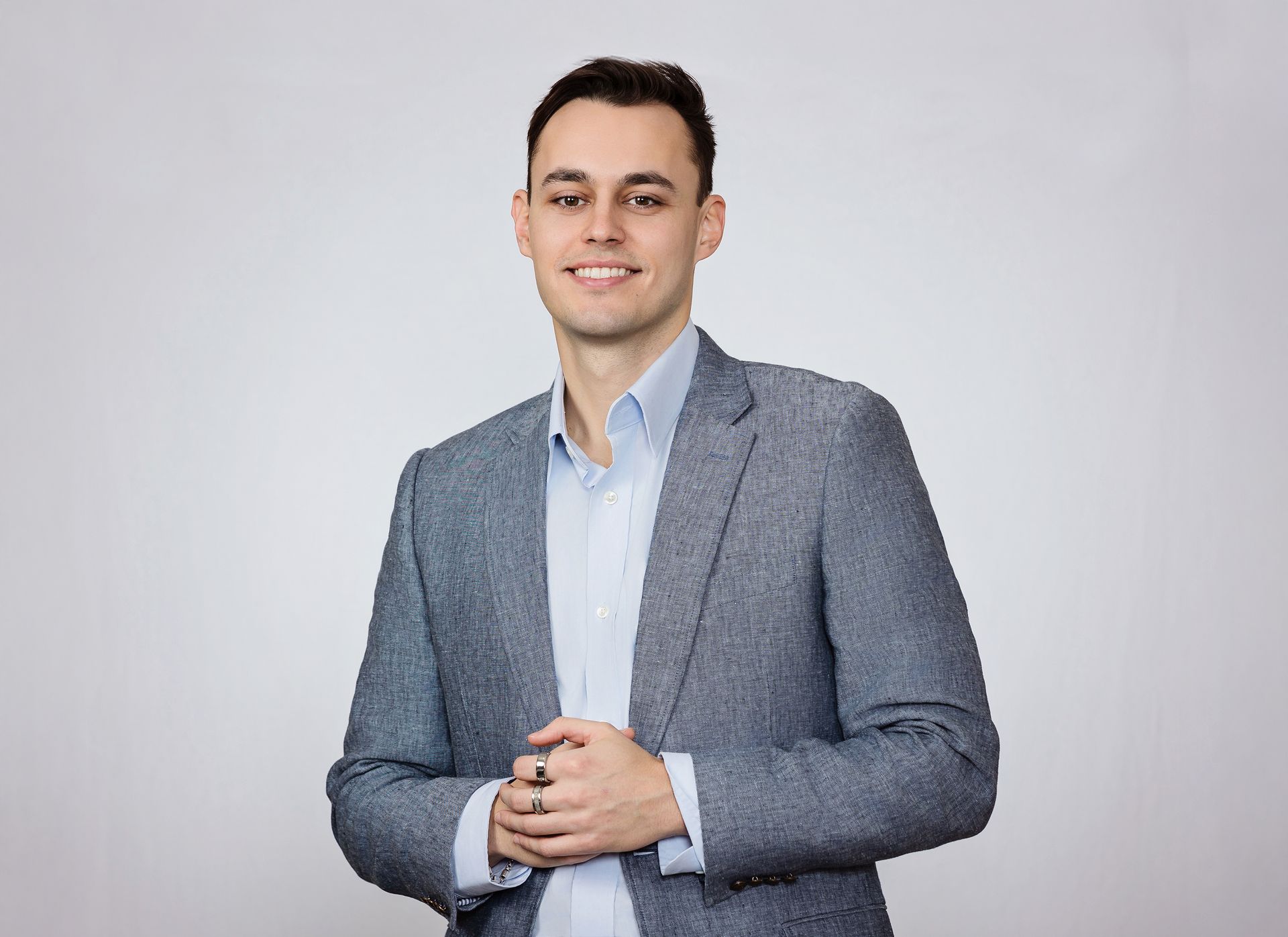 A man in a suit is standing with his hands folded and smiling.