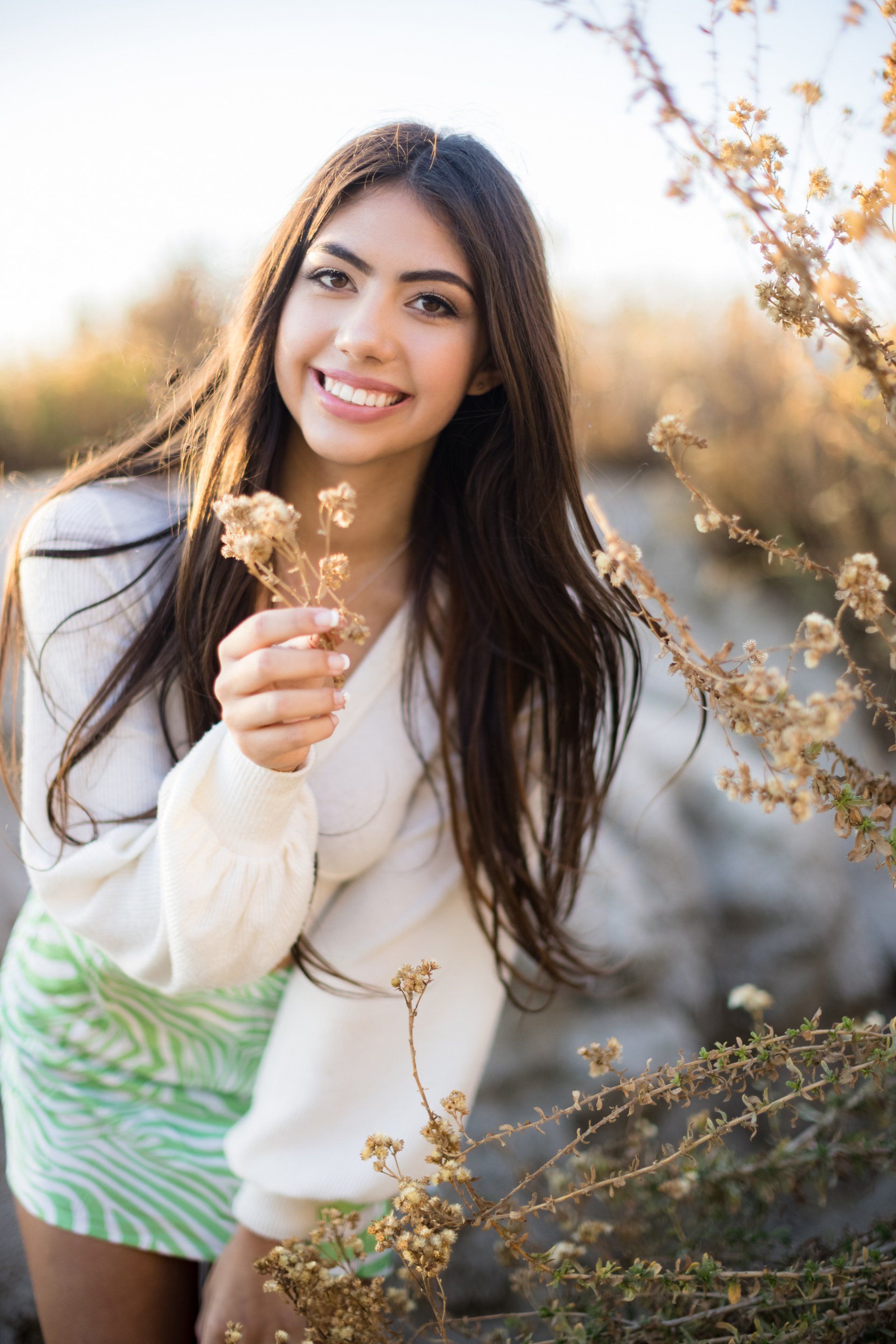 A woman in a white sweater and green skirt is holding a flower and smiling.