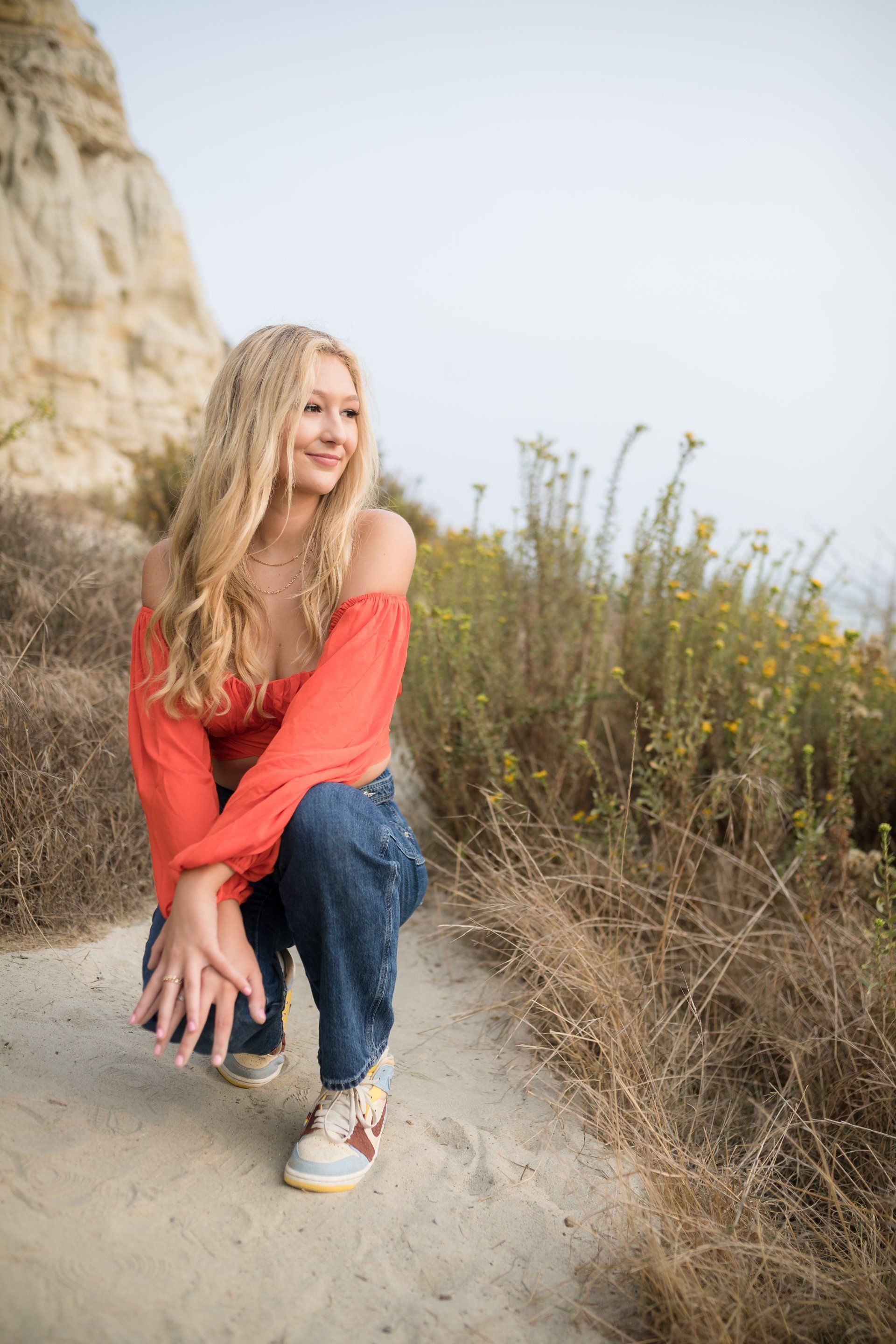 A woman in a red top and jeans is squatting down on a dirt path.