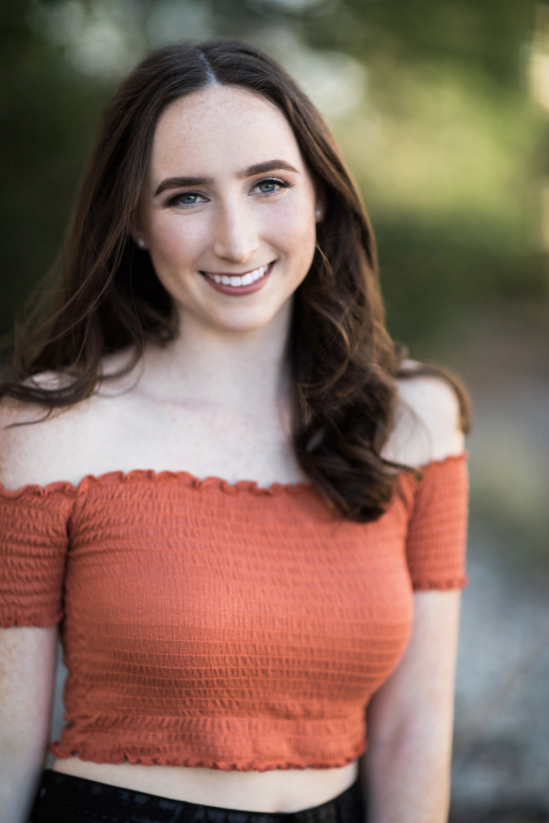 A young woman in an orange off the shoulder top is smiling for the camera.