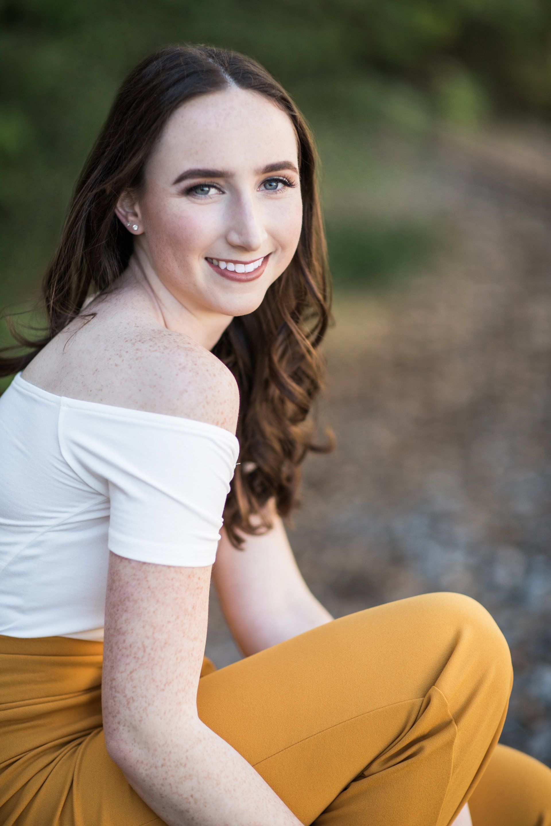 A young woman is sitting on the ground wearing a white off the shoulder top and yellow pants.