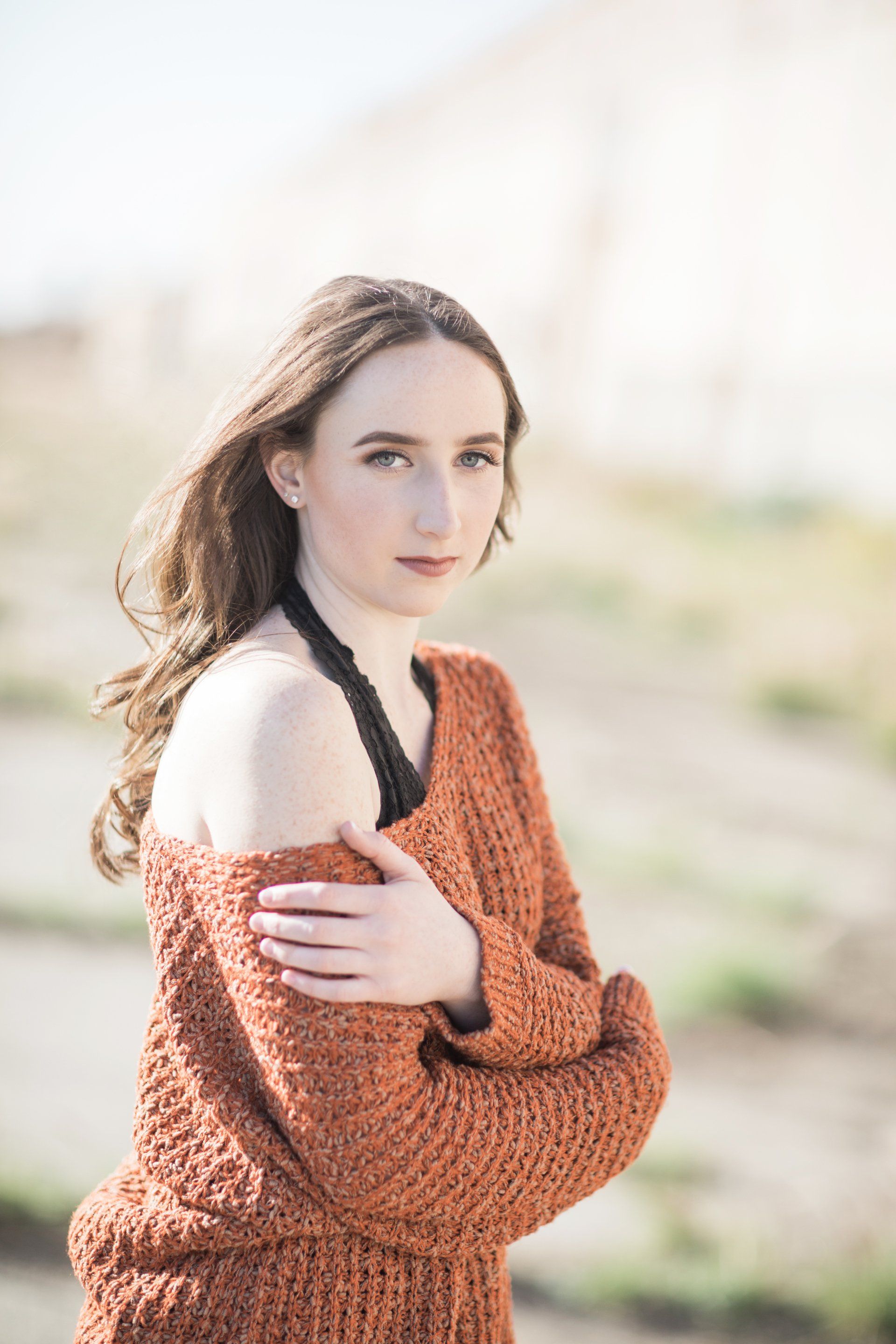 A woman in an orange sweater is standing in a field with her arms crossed.