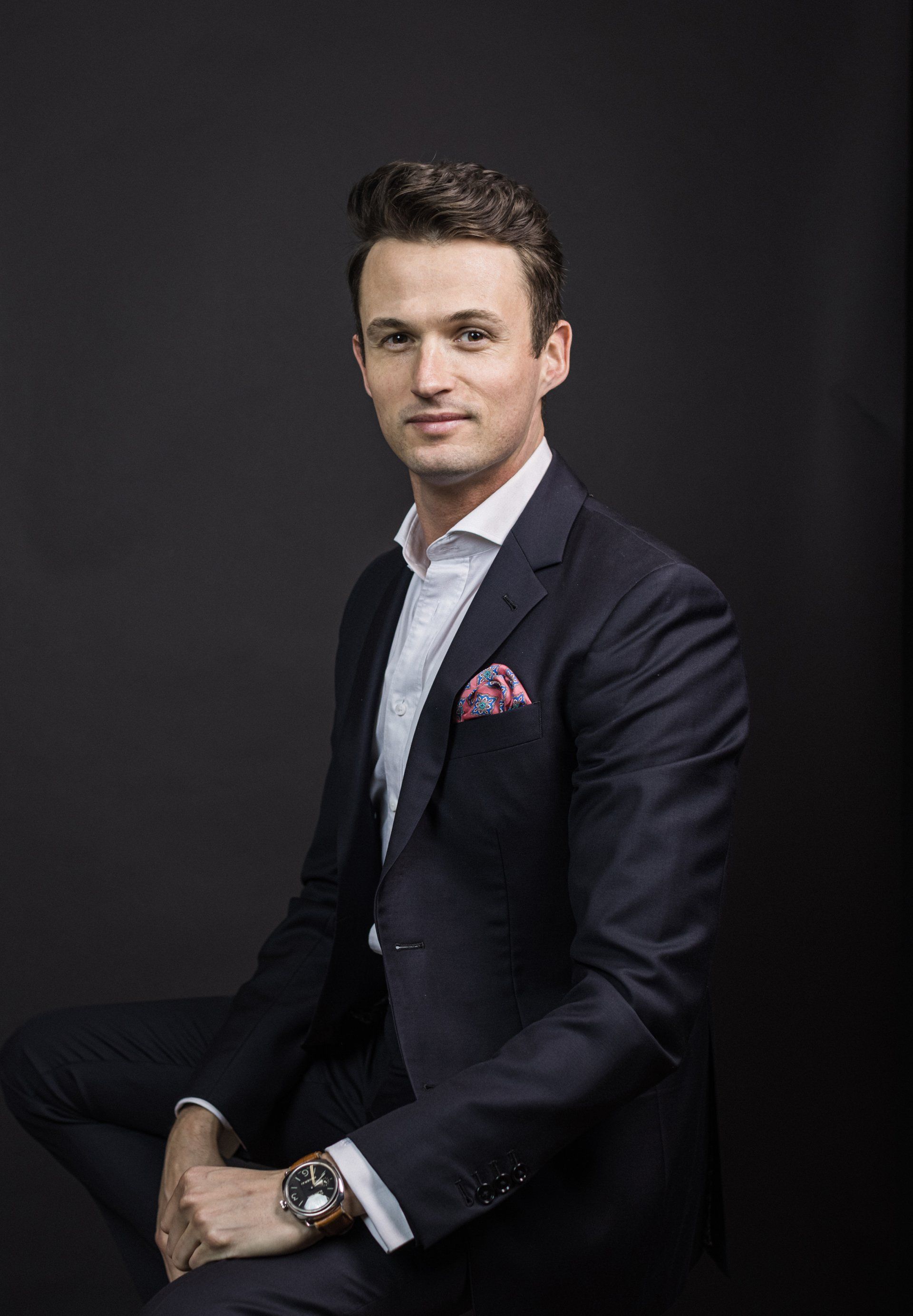 A man in a suit and tie is sitting down and wearing a watch.