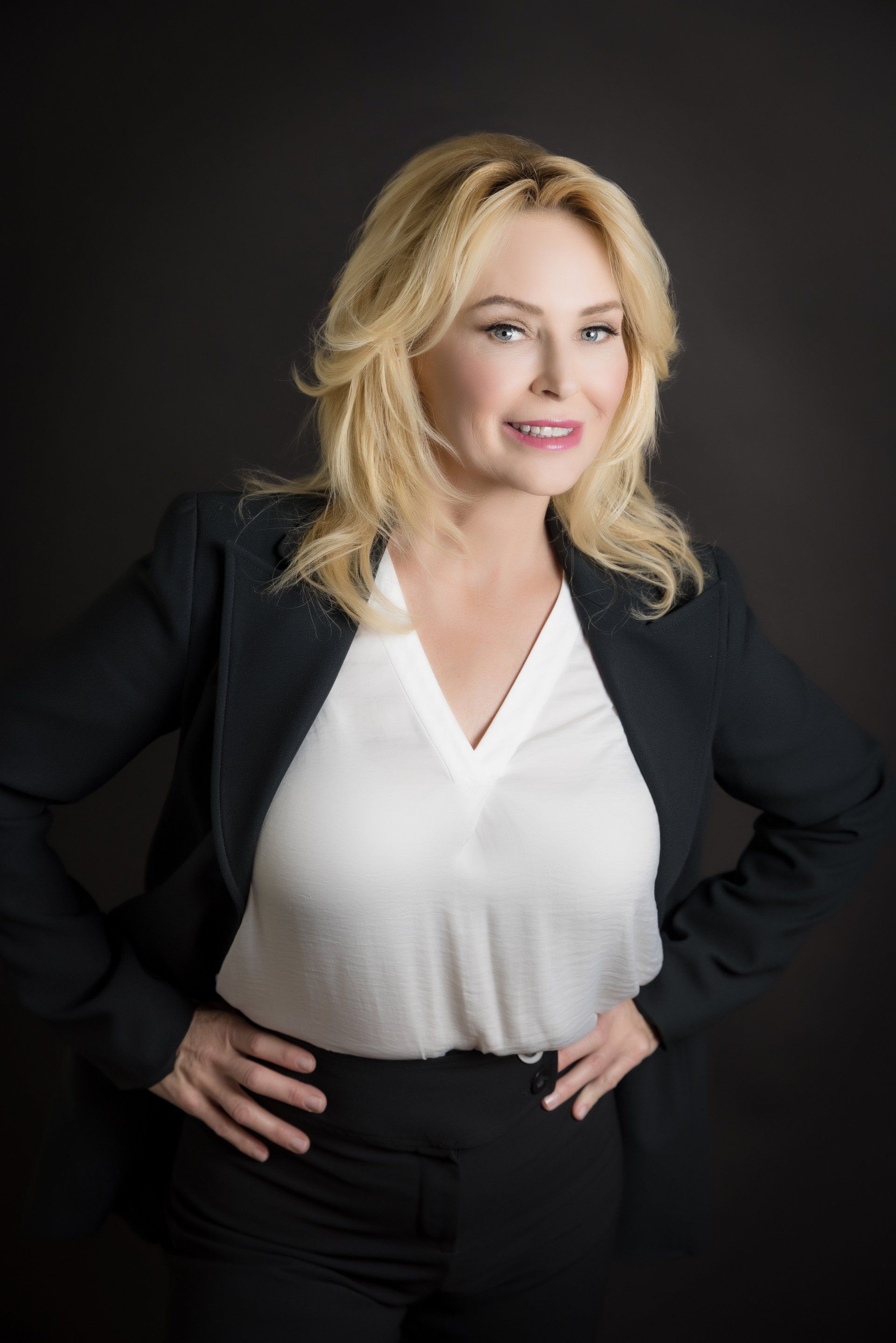 A woman in a white shirt and black jacket is standing with her hands on her hips.