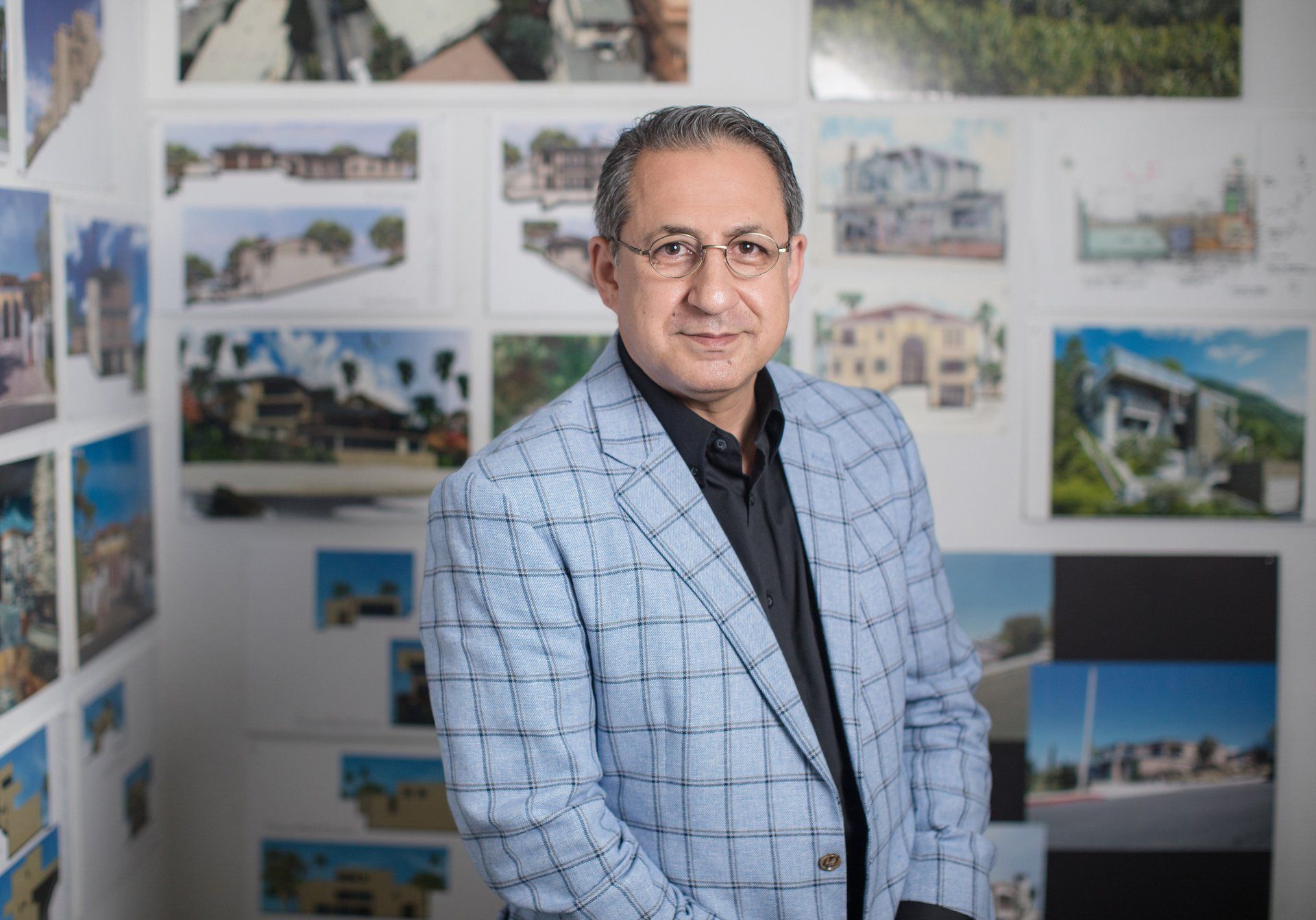 A man in a suit and glasses is standing in front of a wall of pictures.