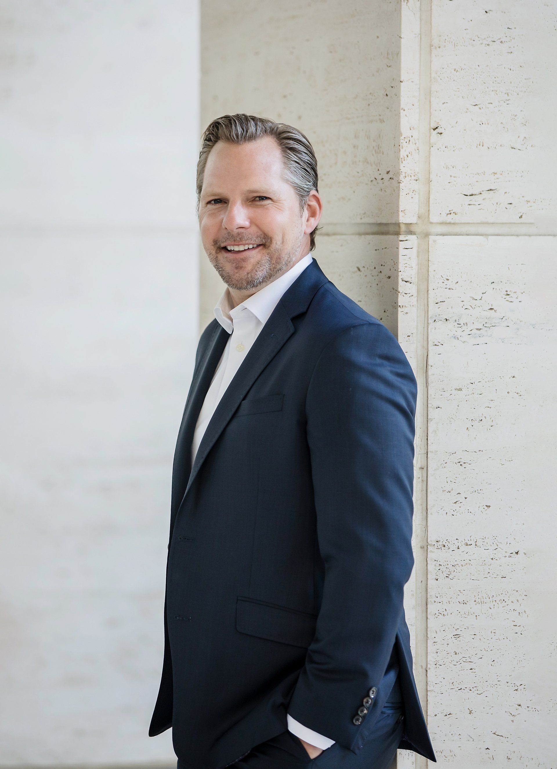 A man in a suit is leaning against a wall with his hands in his pockets.