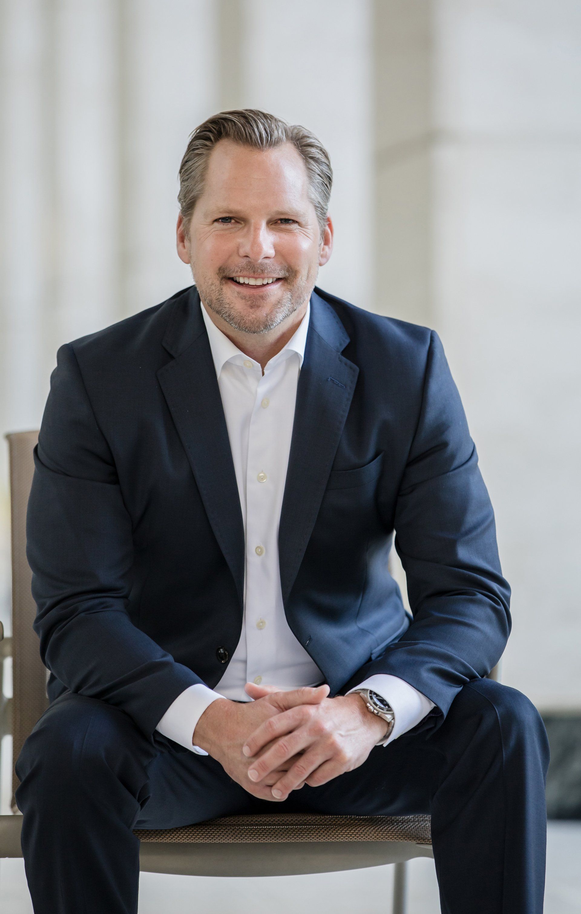A man in a suit is sitting on a chair with his hands folded and smiling.