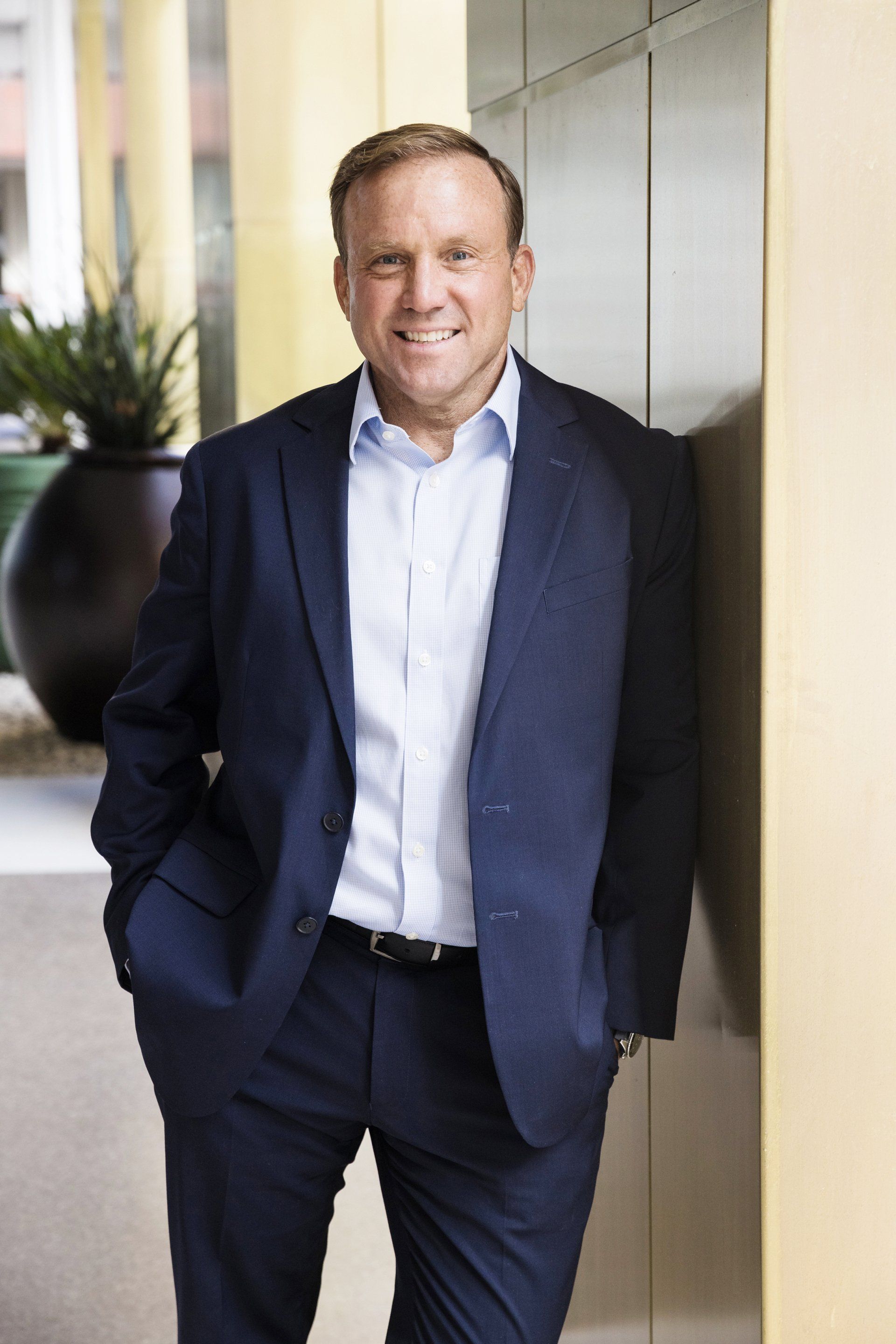 A man in a suit is leaning against a wall with his hands in his pockets.