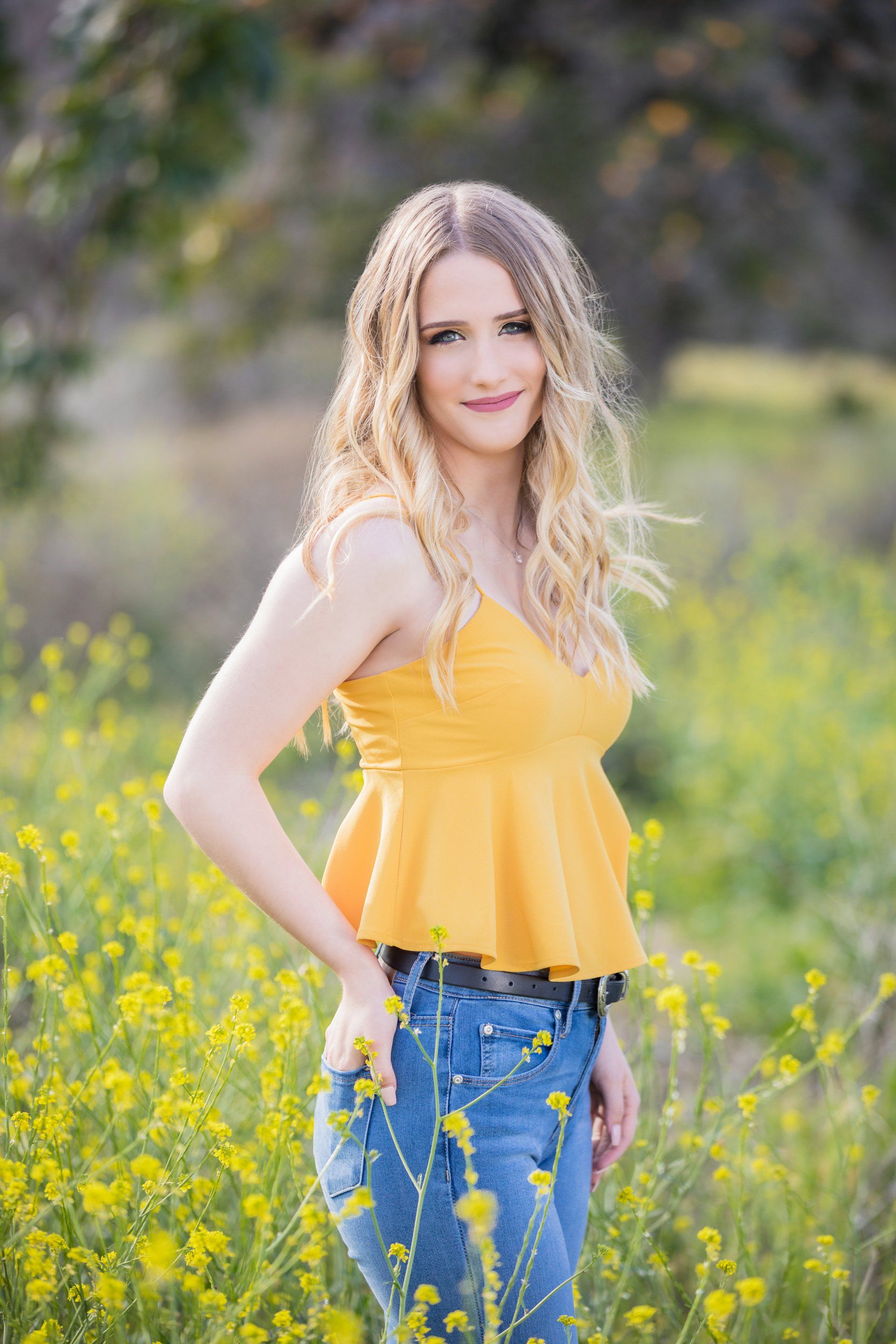 A woman in a yellow tank top and blue jeans is standing in a field of yellow flowers.