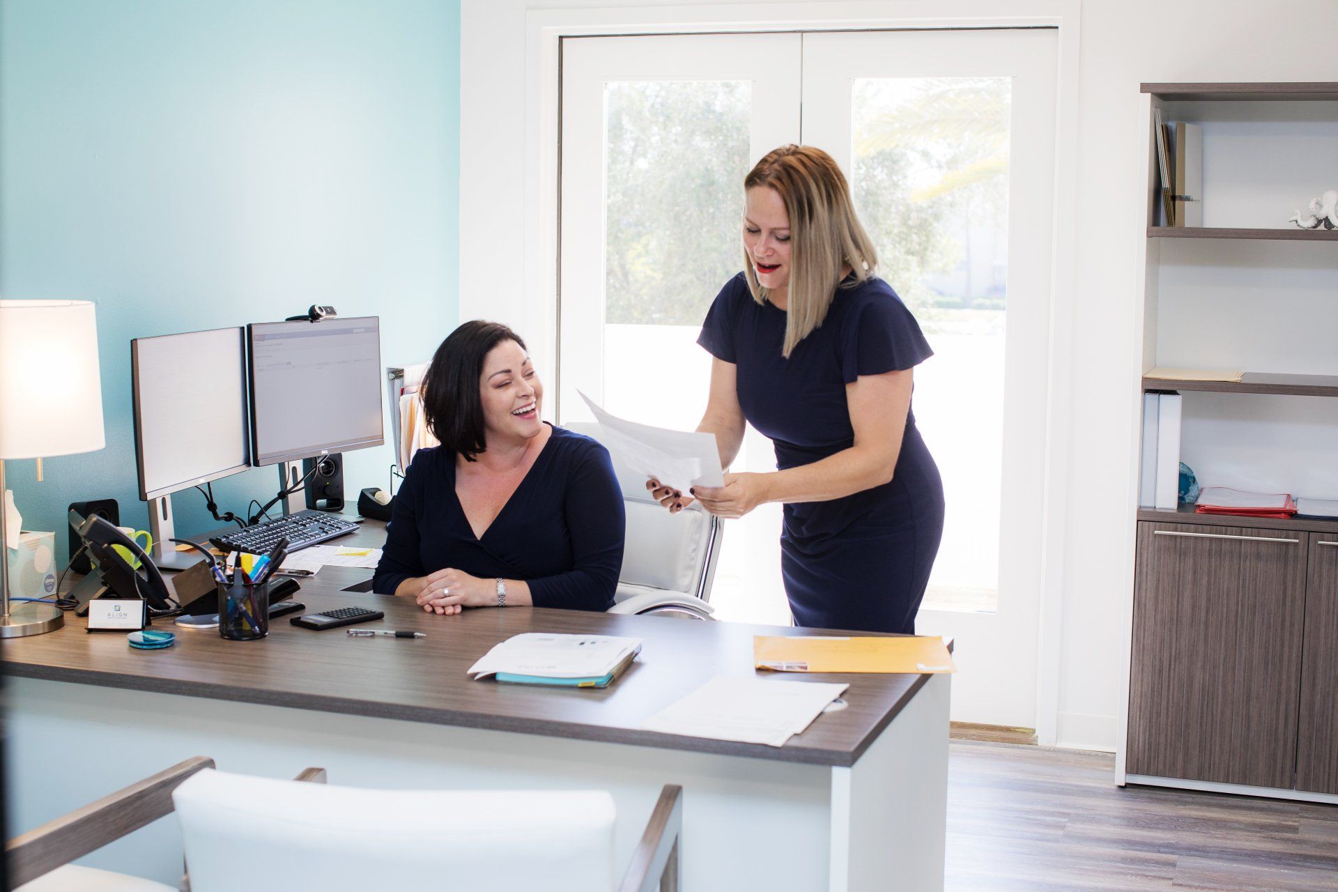 Two women are standing next to each other at a desk in an office.