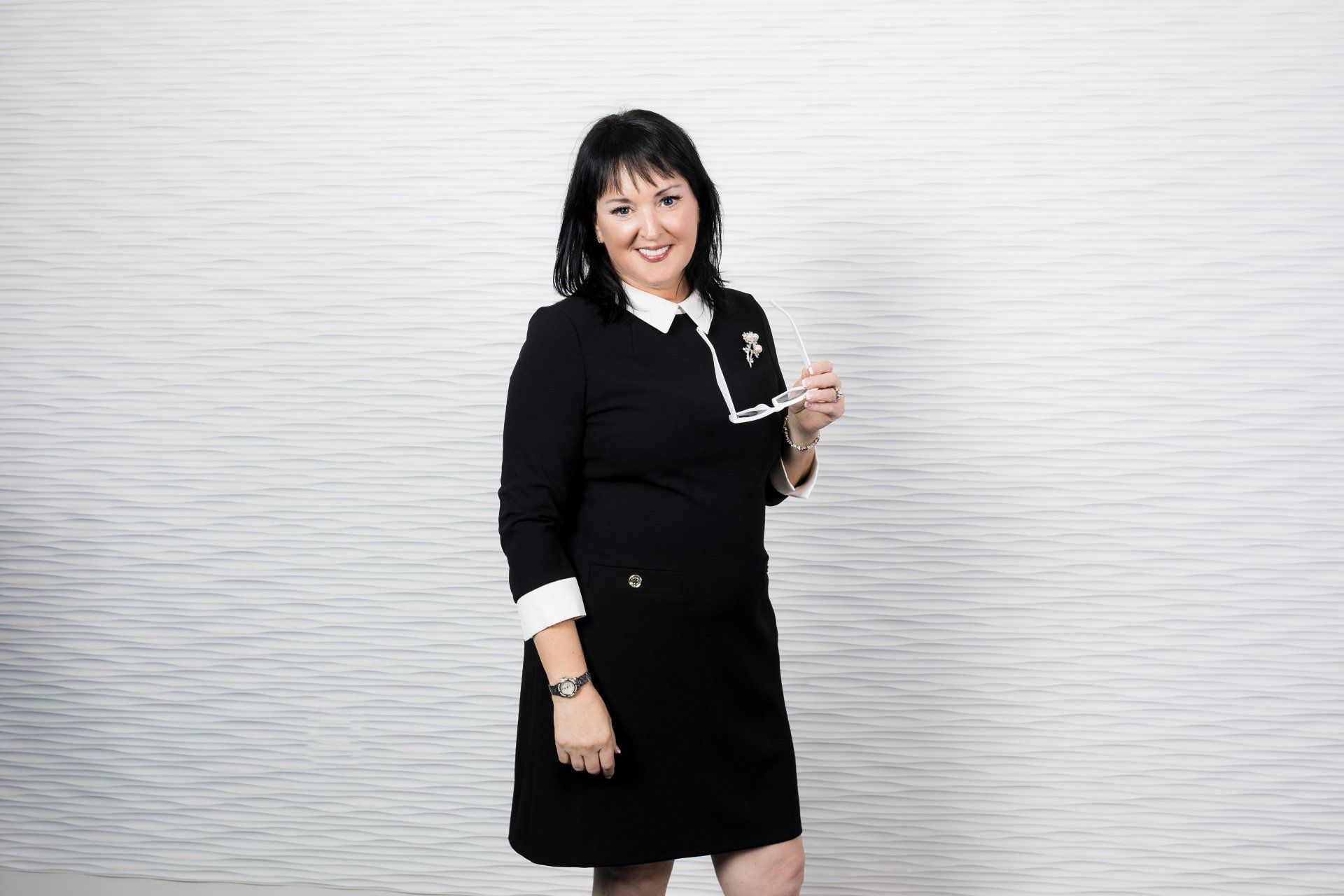 A woman in a black dress is standing in front of a white wall holding a pair of glasses.