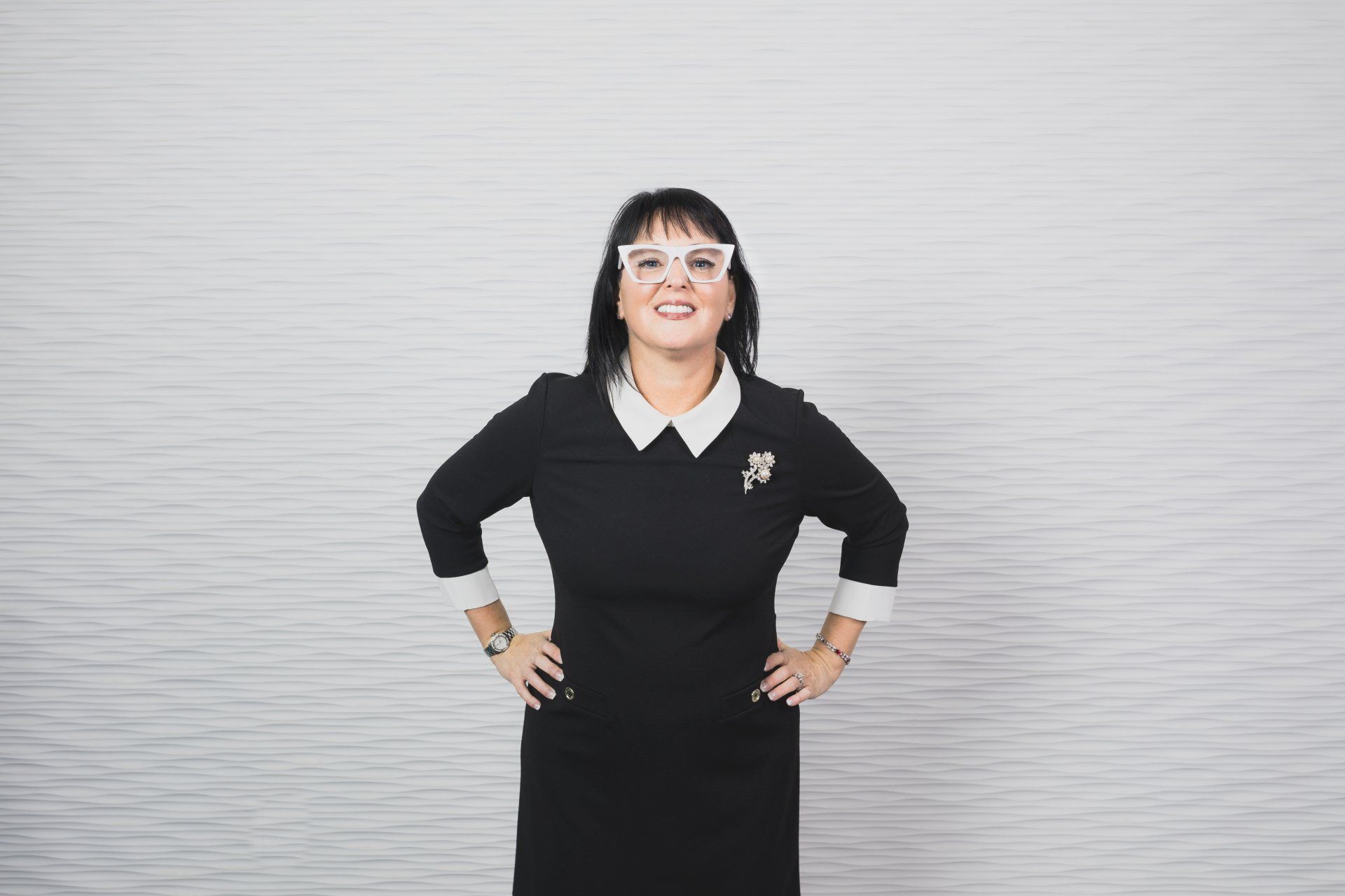 A woman in a black dress and white glasses is standing with her hands on her hips.