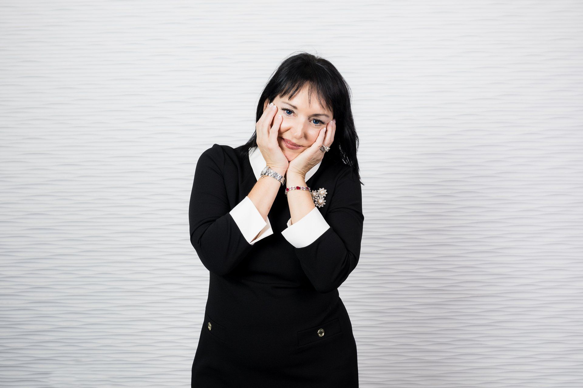 A woman in a black dress is standing in front of a white wall with her hands on her face.