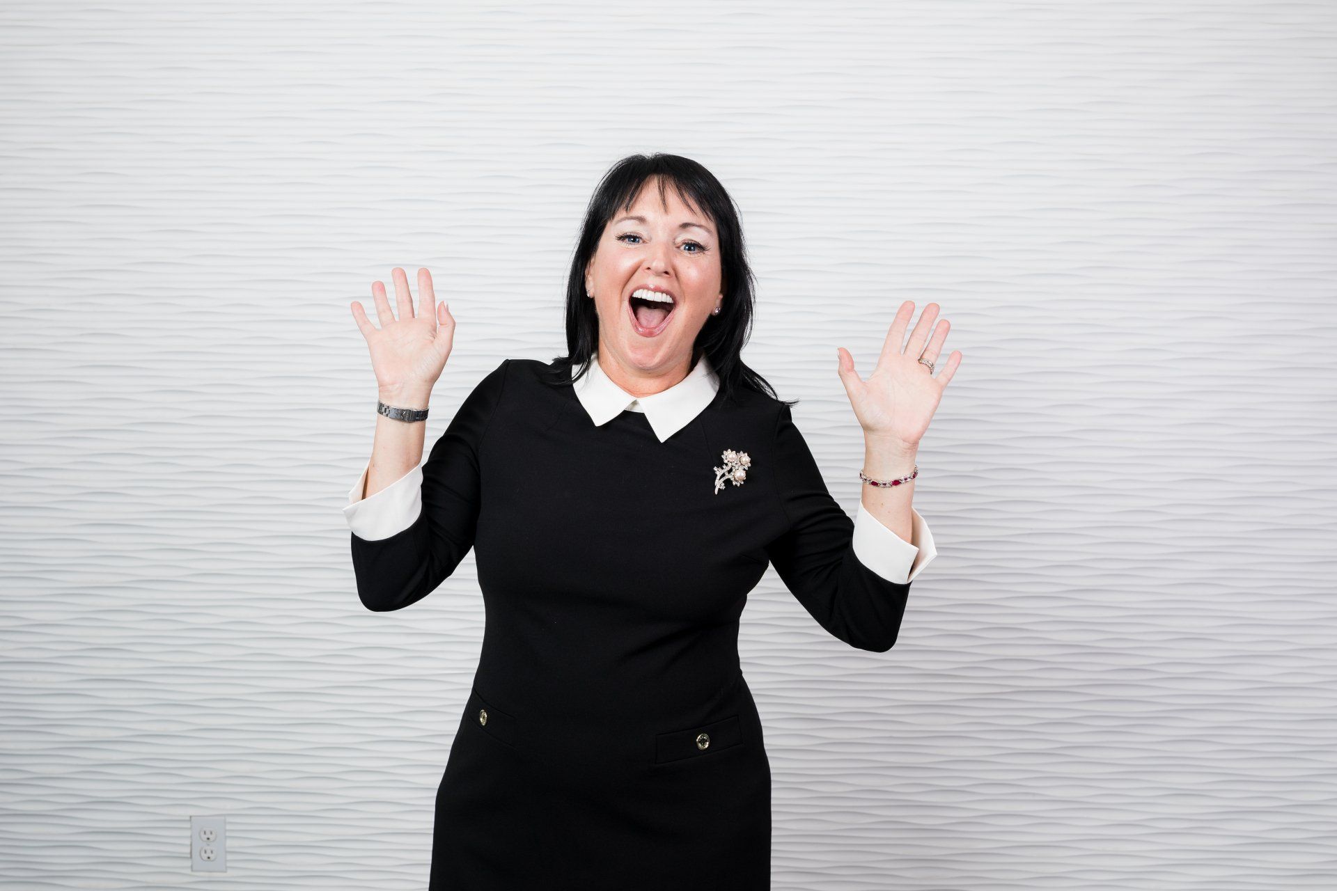 A woman in a black dress is standing in front of a white wall with her hands in the air.