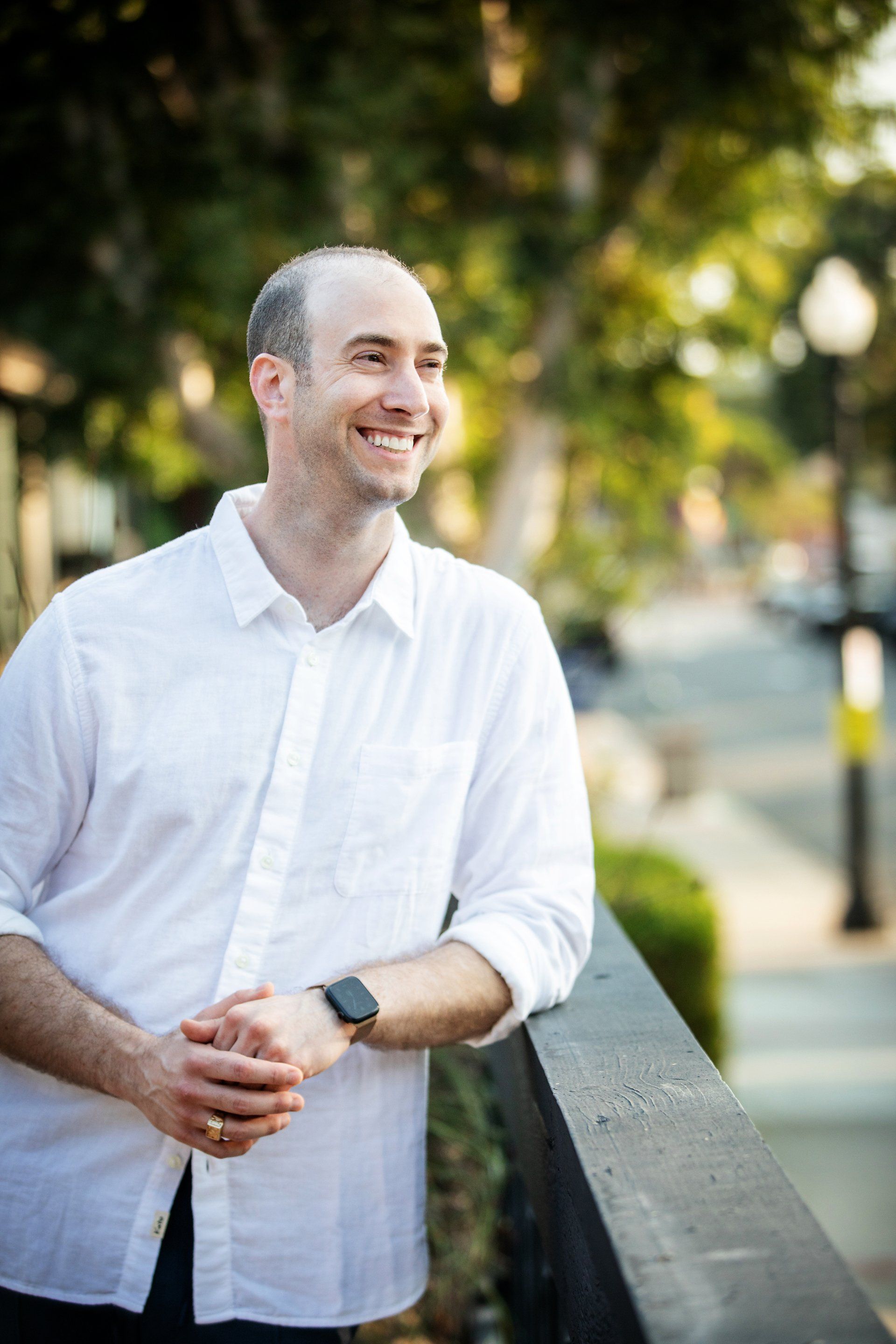 A man in a white shirt is leaning against a railing and smiling.