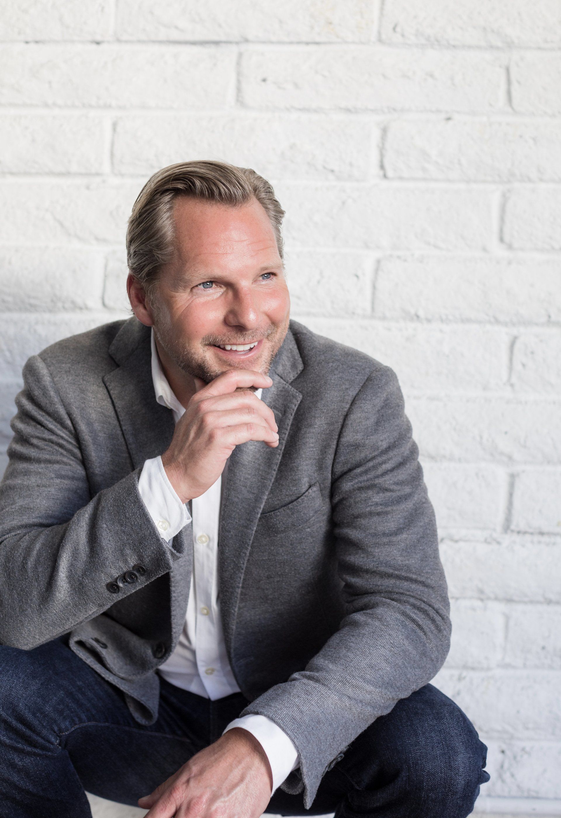 A man in a suit is sitting in front of a white brick wall.