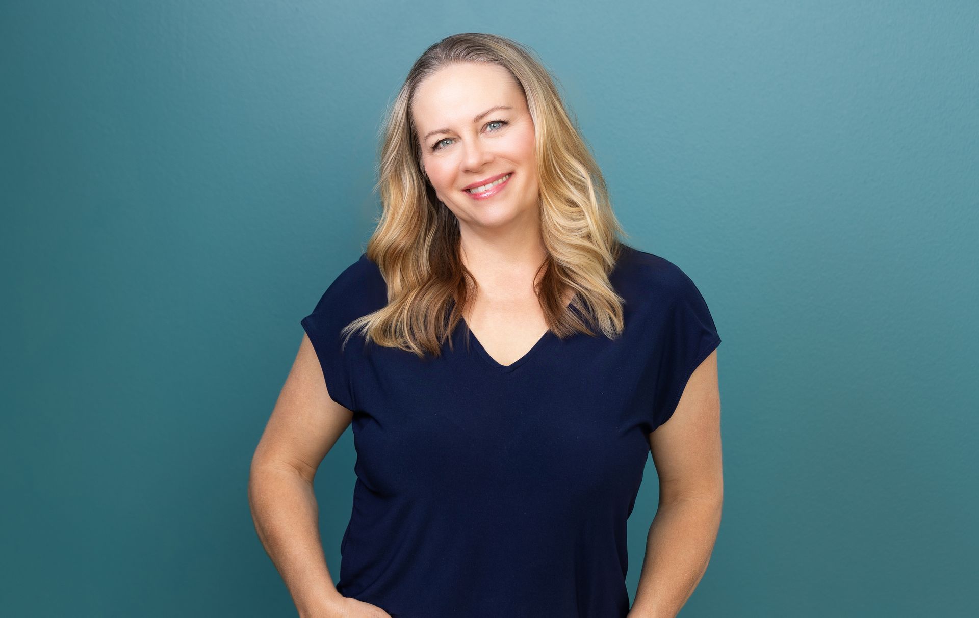 A woman in a blue shirt is standing in front of a blue wall and smiling.