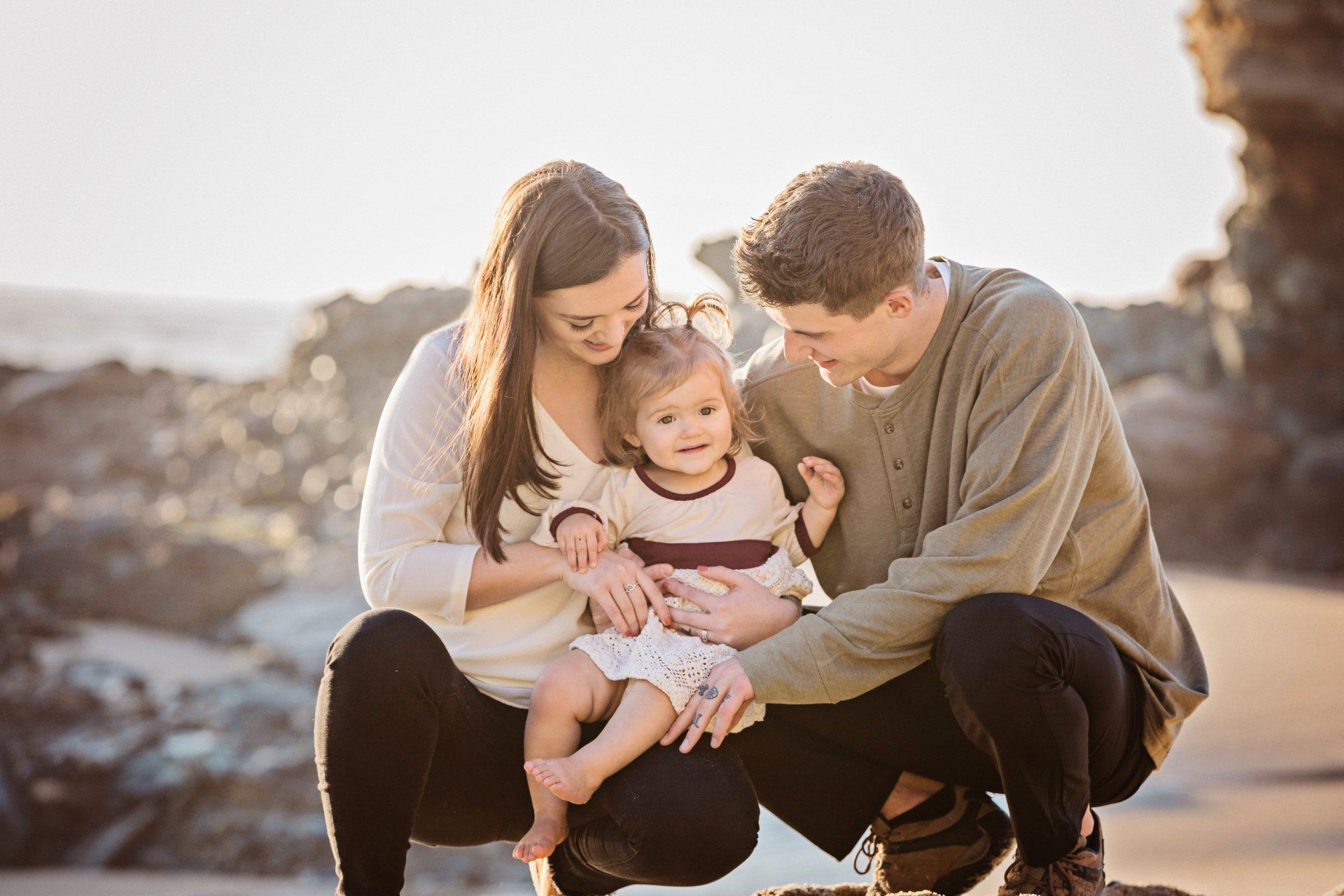 A young family, captured by an Orange County family photographer from Blue Sky’s Studio, enjoys a serene moment together on a sunlit Laguna Beach. The parents, dressed in neutral tones, lovingly embrace their smiling toddler, creating a warm and intimate scene. The rocky beach setting, bathed in golden light, highlights the natural beauty typical of Orange County family portraits, making this a memorable and heartwarming image that embodies the essence of coastal photography.