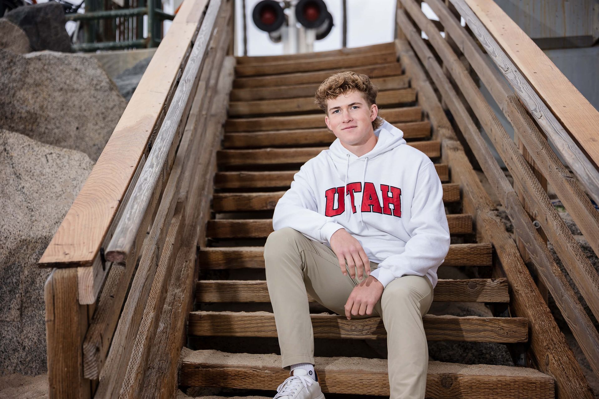 High school senior boy wearing a University of Utah hoodie during his senior portrait session on wooden stairs at the beach in Orange County, photographed by Blue Sky’s Studio