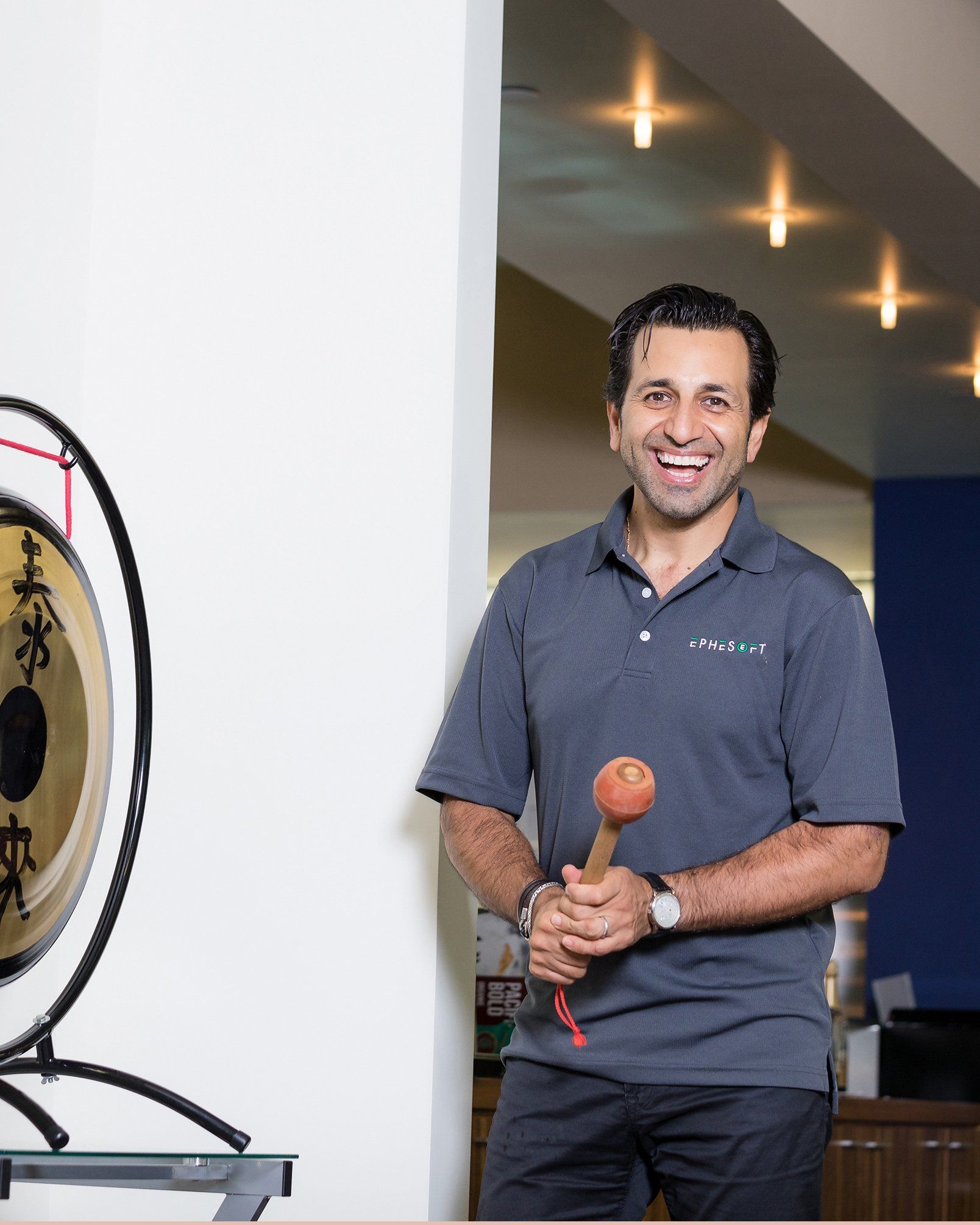 A man in a grey shirt is smiling in front of a gong