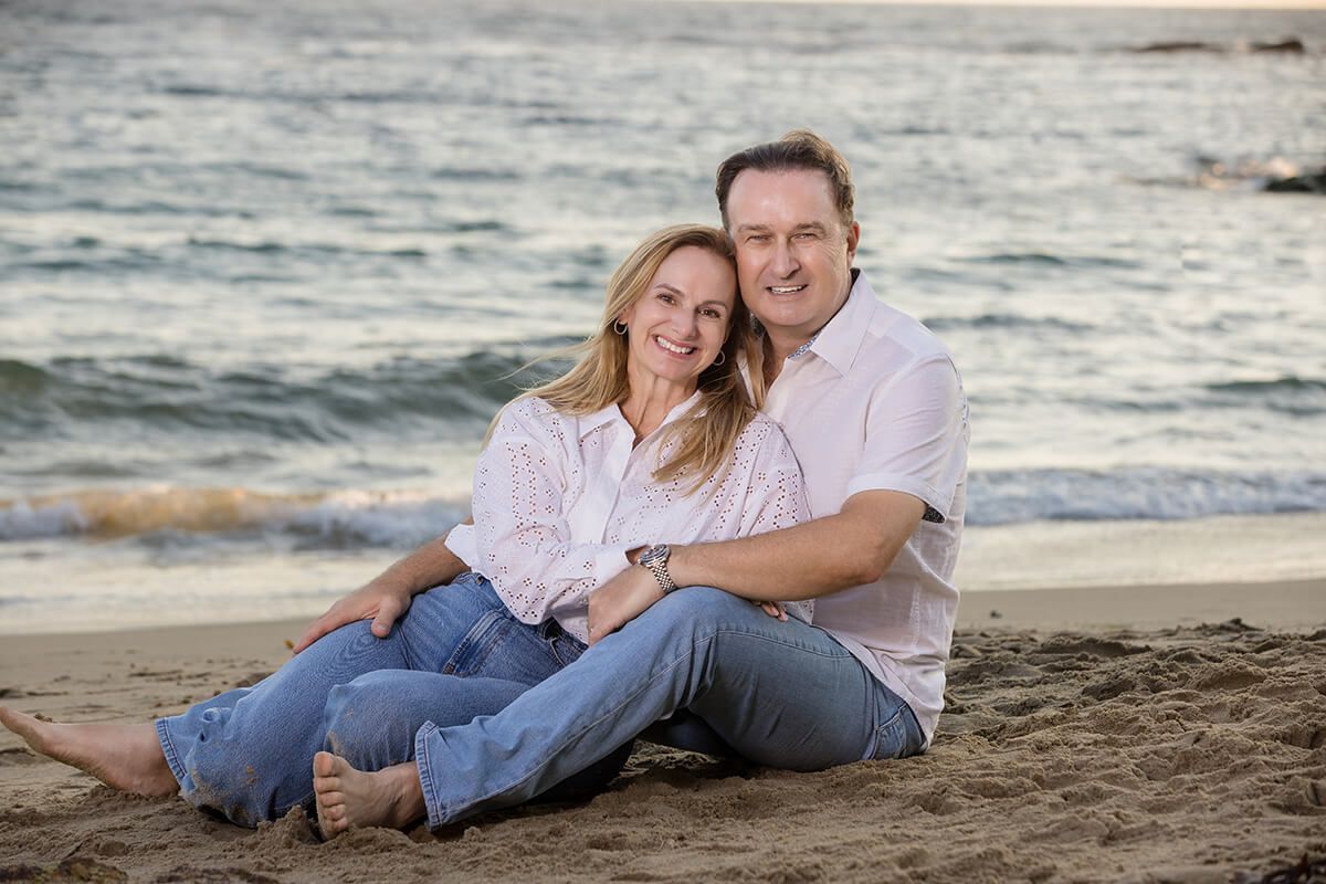 Couple sitting on the sand at Laguna Beach during sunset, smiling and embracing—captured by Orange County family photographer.