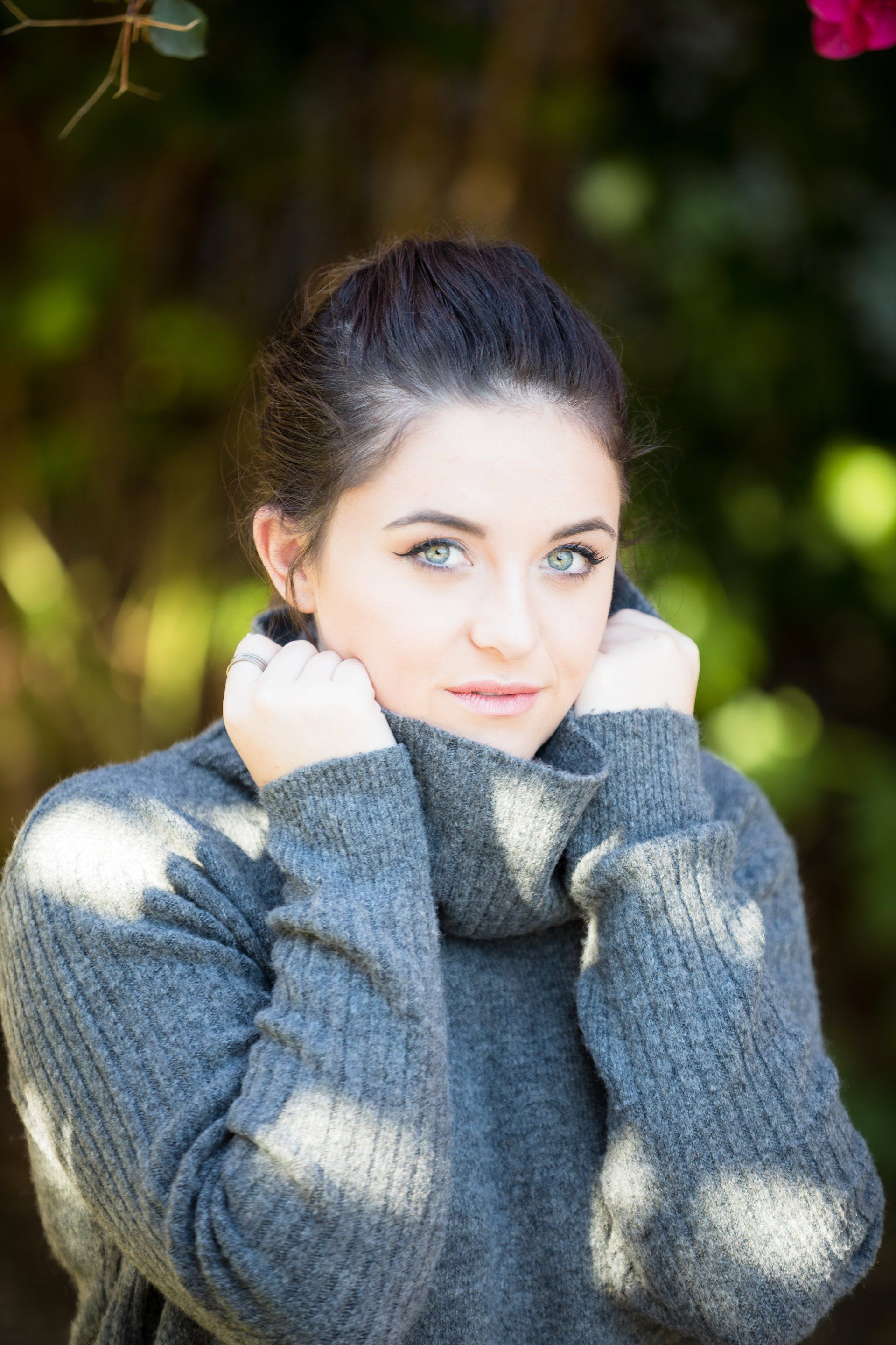 A young woman wearing a turtleneck sweater is standing in front of a bush.