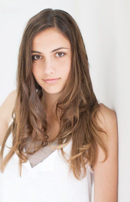 A young woman with long hair is leaning against a white wall.