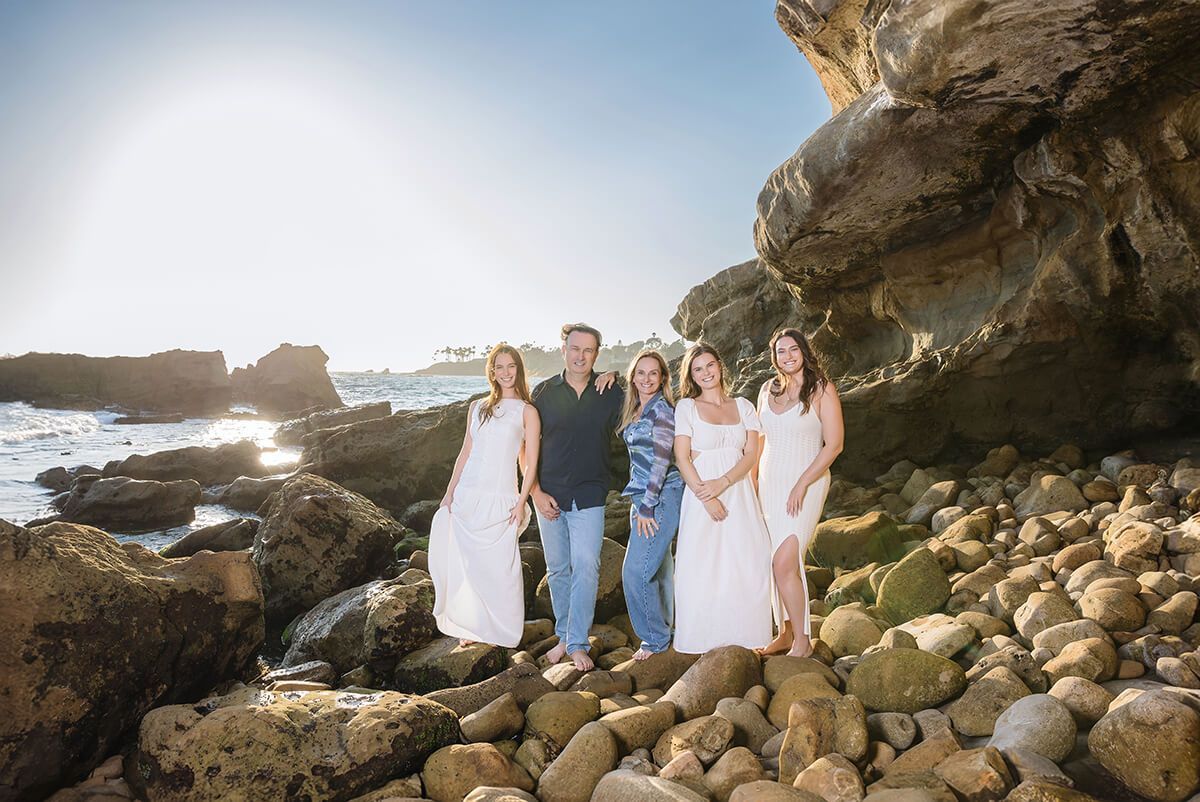 Family standing on rocky beach in Laguna Beach during golden hour, wearing coordinated white and blue outfits—captured by Orange County family photographer.