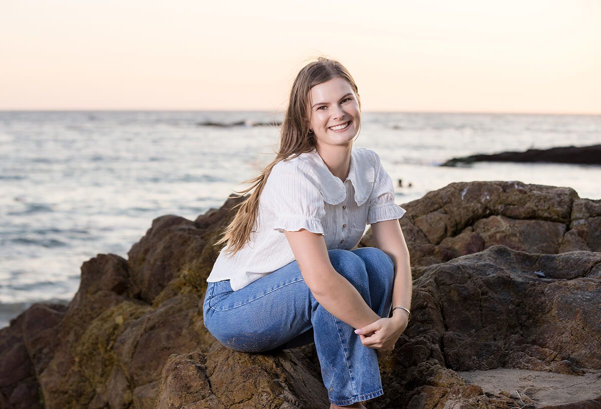 Golden hour beach portrait of a young woman sitting on coastal rocks in Laguna Beach, dressed in a white blouse and jeans, photographed by Orange County family and senior photographer Blue Sky’s Studio.
