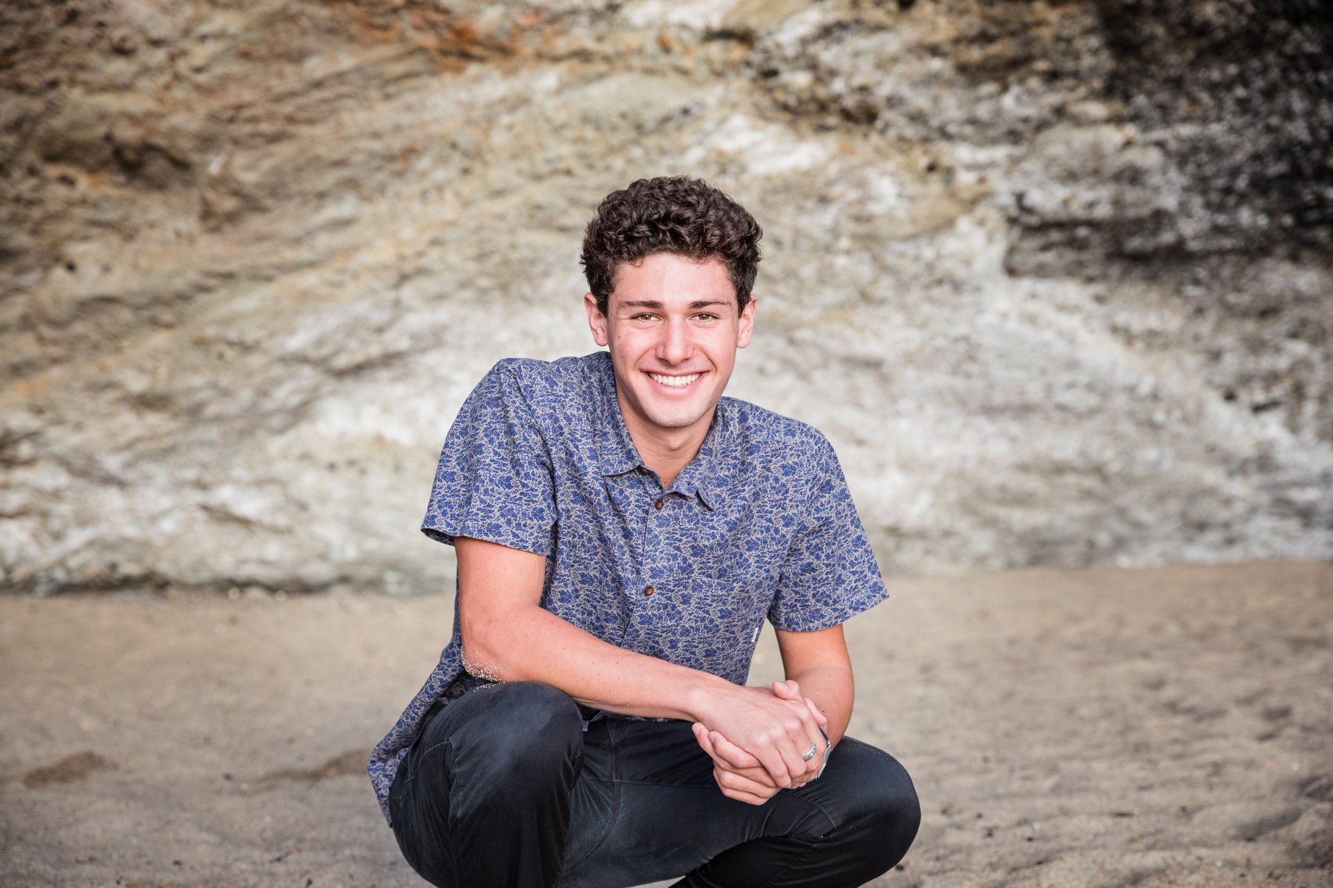 A young man is squatting down on the beach and smiling.