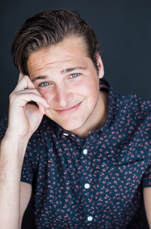 A young man in a blue shirt is smiling with his hand on his face.