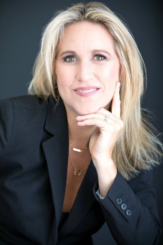 A woman in a black suit is smiling with her hand on her chin.