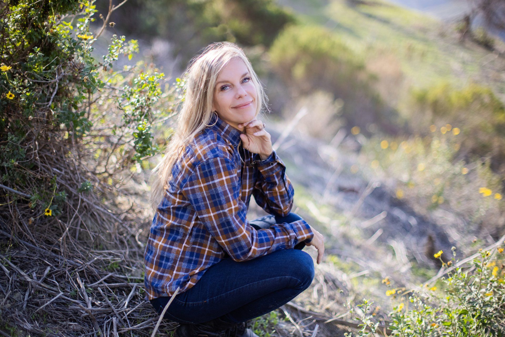A woman in a plaid shirt is squatting down in the grass.