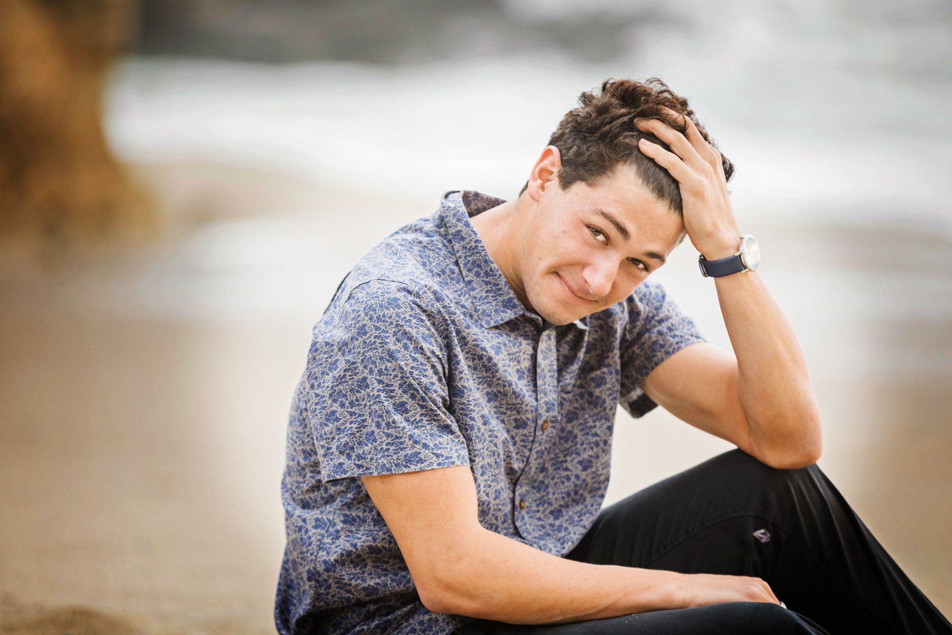 A young man is sitting on the beach with his hand on his head.