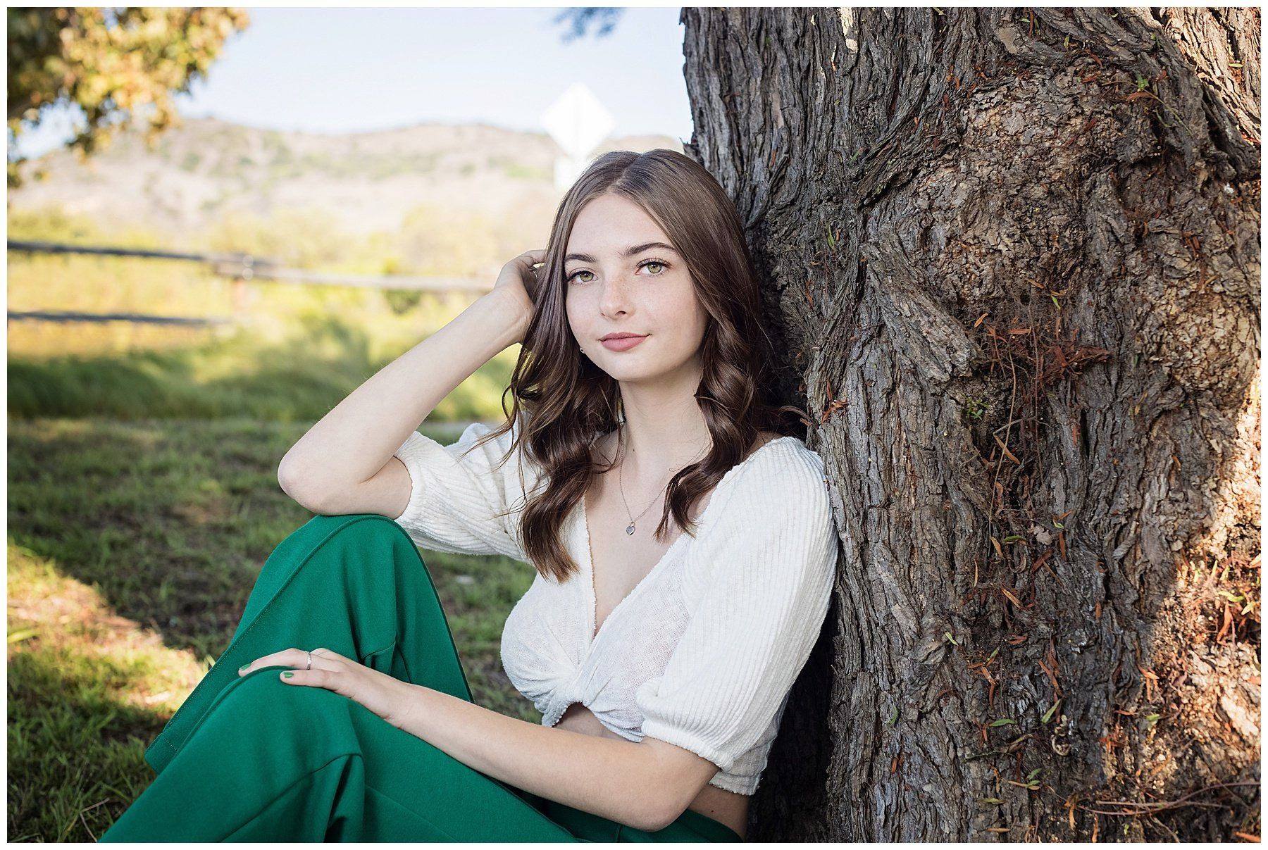 A woman in a white top and green pants is leaning against a tree.