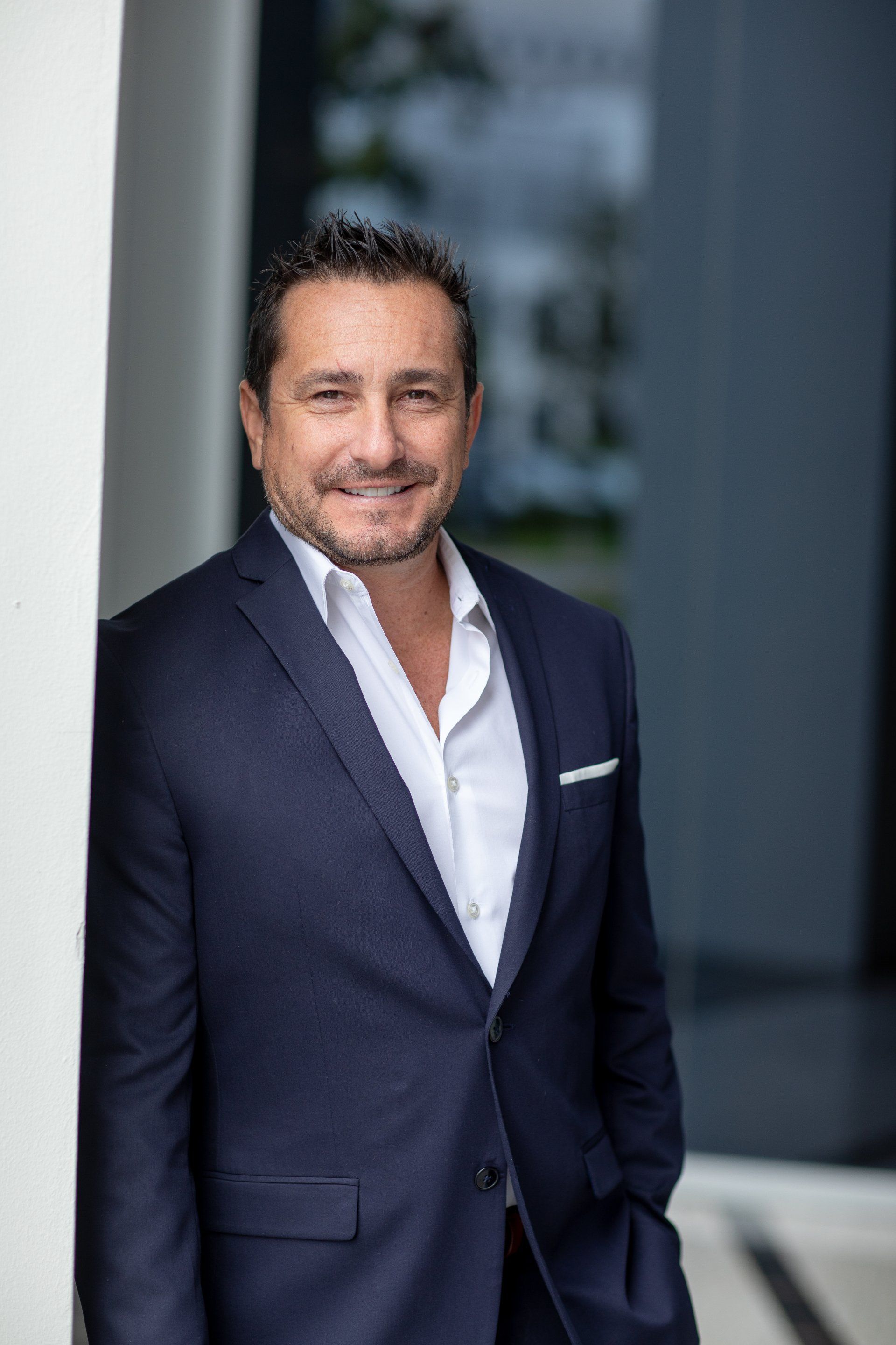 A man in a suit and white shirt is leaning against a wall.