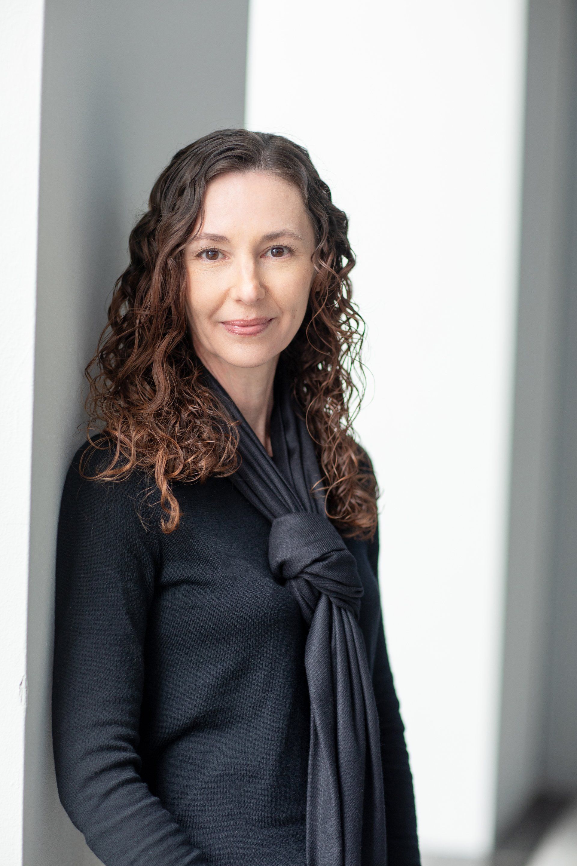 A woman in a black sweater and scarf is leaning against a wall.