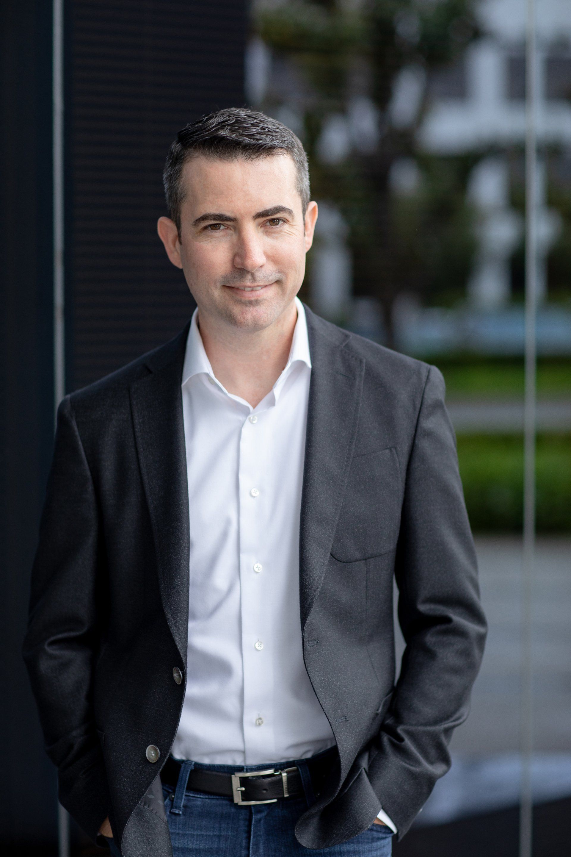 A man in a suit and white shirt is standing in front of a building with his hands in his pockets.