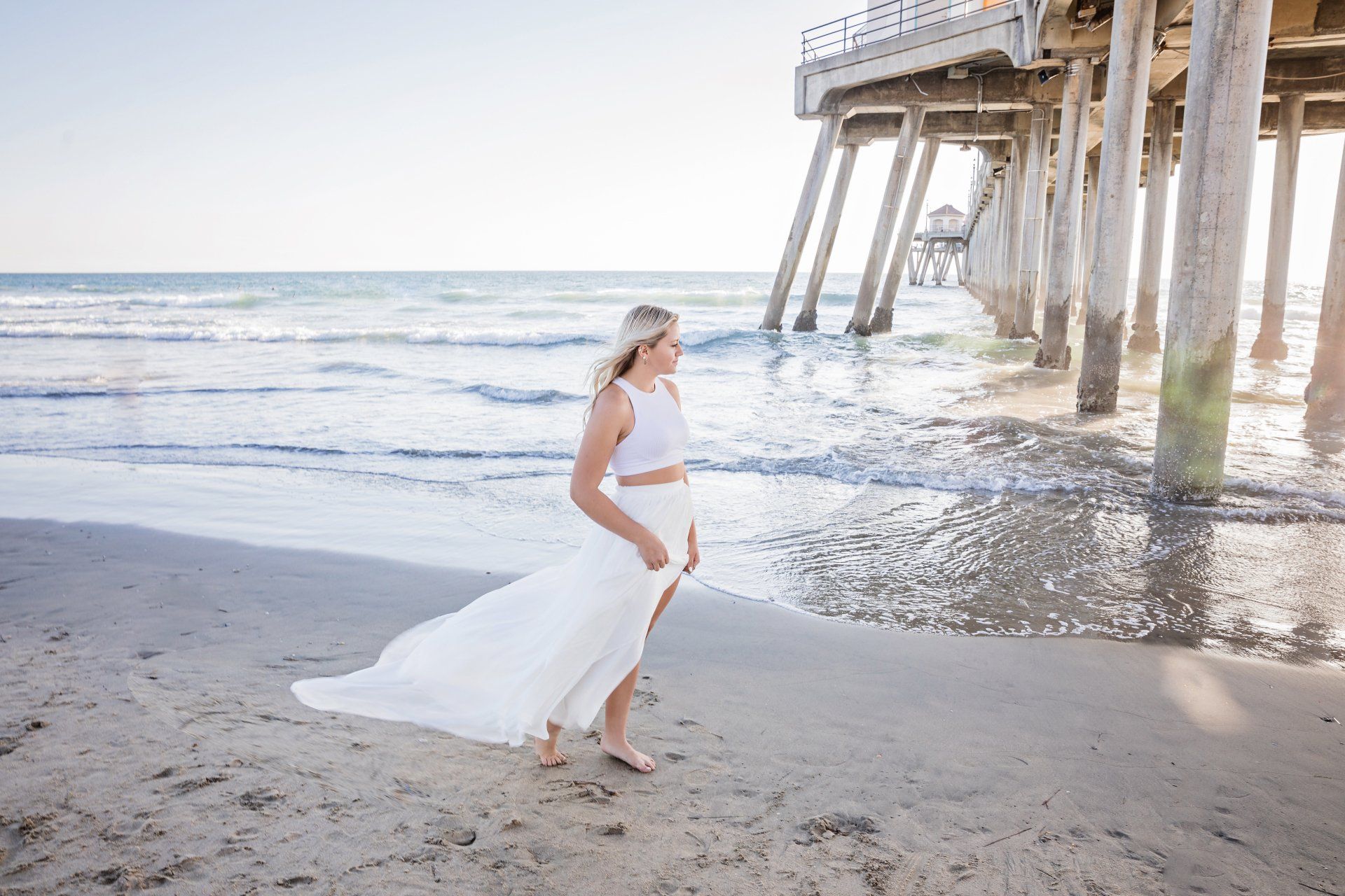A woman in a white dress is standing on the beach under a pier.