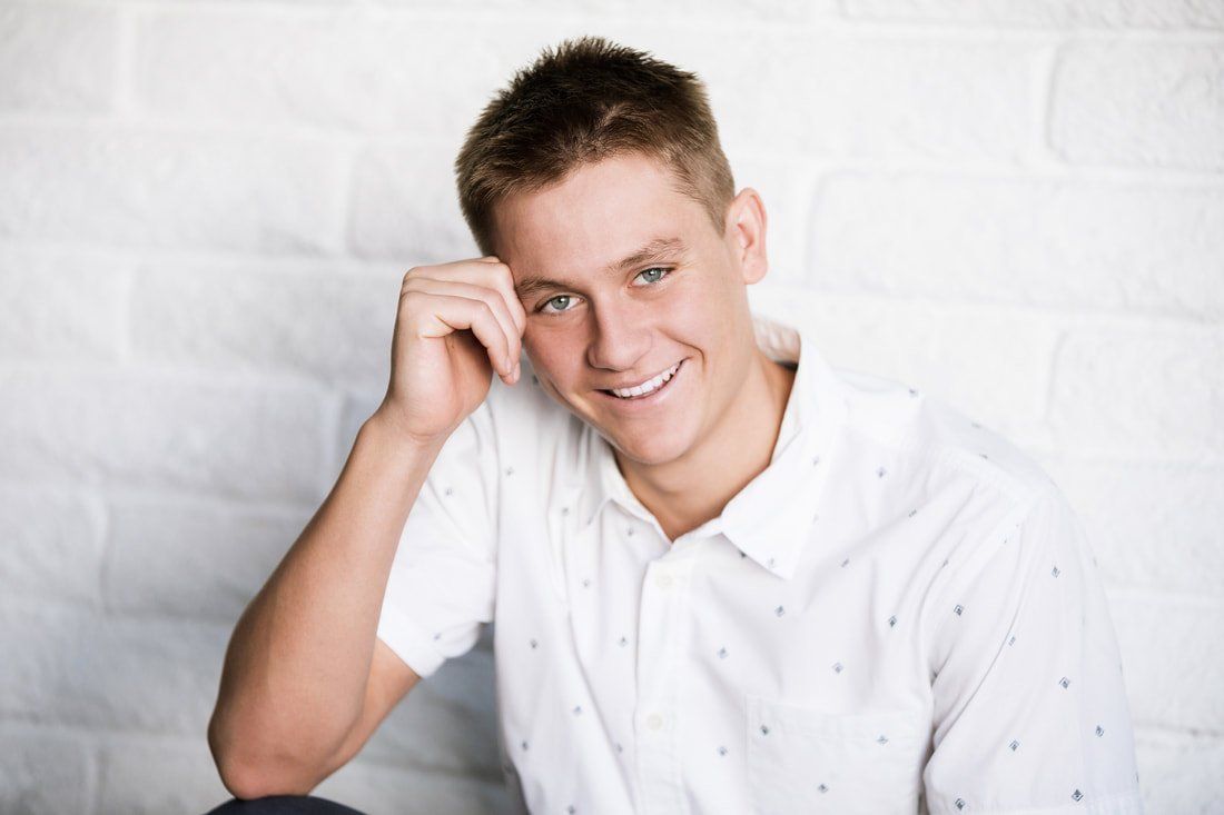 A young man in a white shirt is sitting in front of a white brick wall.