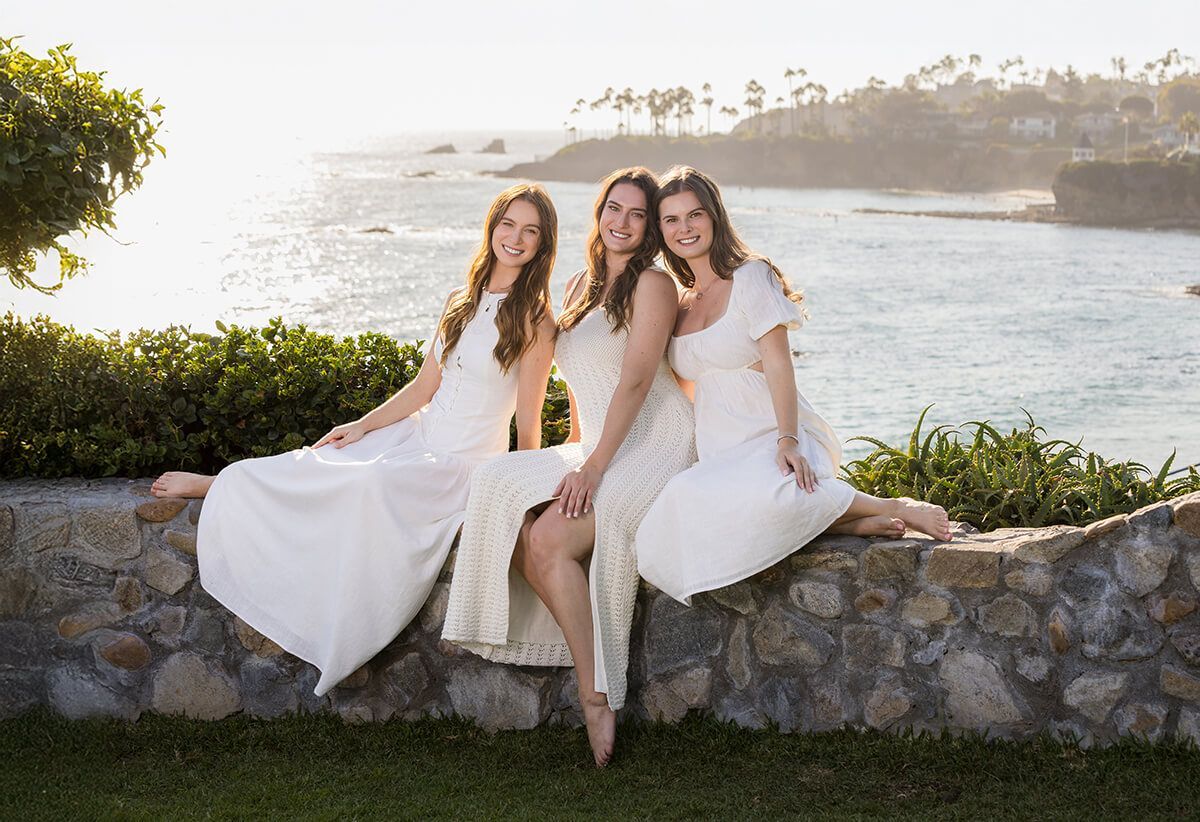 Three women in white dresses sitting on a stone wall overlooking the ocean at sunset during a Laguna Beach family portrait session by Orange County family photographer Blue Sky’s Studio.