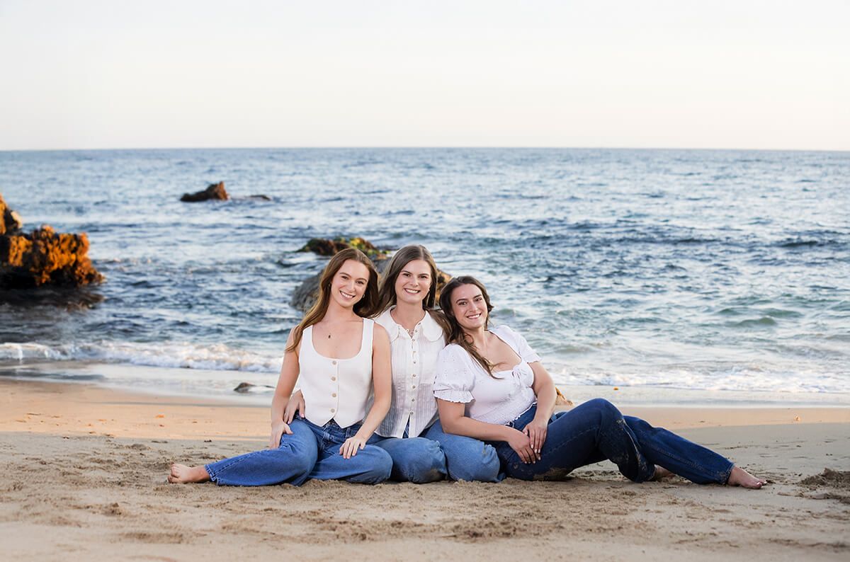 Three sisters sitting on the sand in coordinated white tops and jeans during a sibling beach portrait session in Laguna Beach, captured by Orange County family photographer Blue Sky’s Studio.