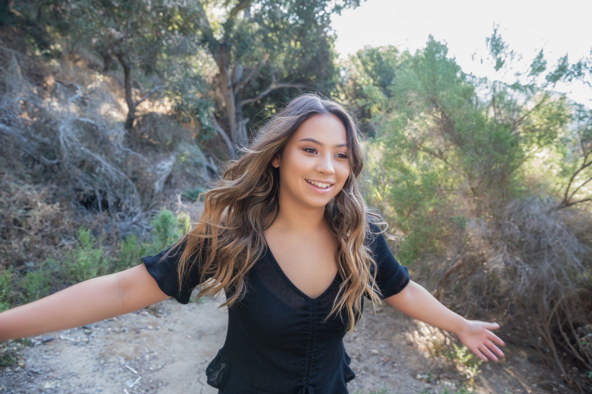 A woman in a black shirt is standing in the woods with her arms outstretched.