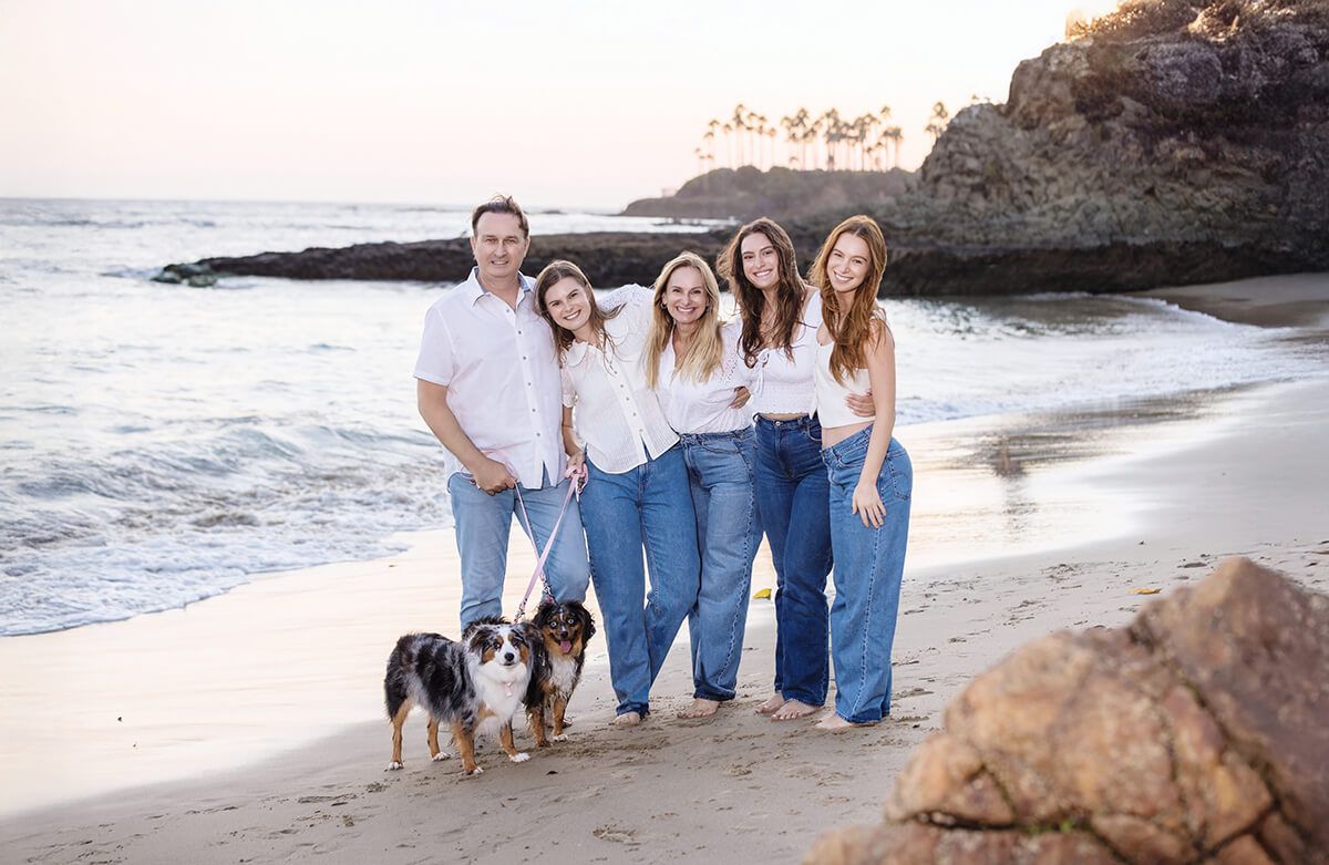 Lifestyle family portrait at sunset on the beach in Laguna Beach with two dogs, featuring parents and three daughters in white tops and denim, photographed by Orange County family photographer Blue Sky’s Studio.