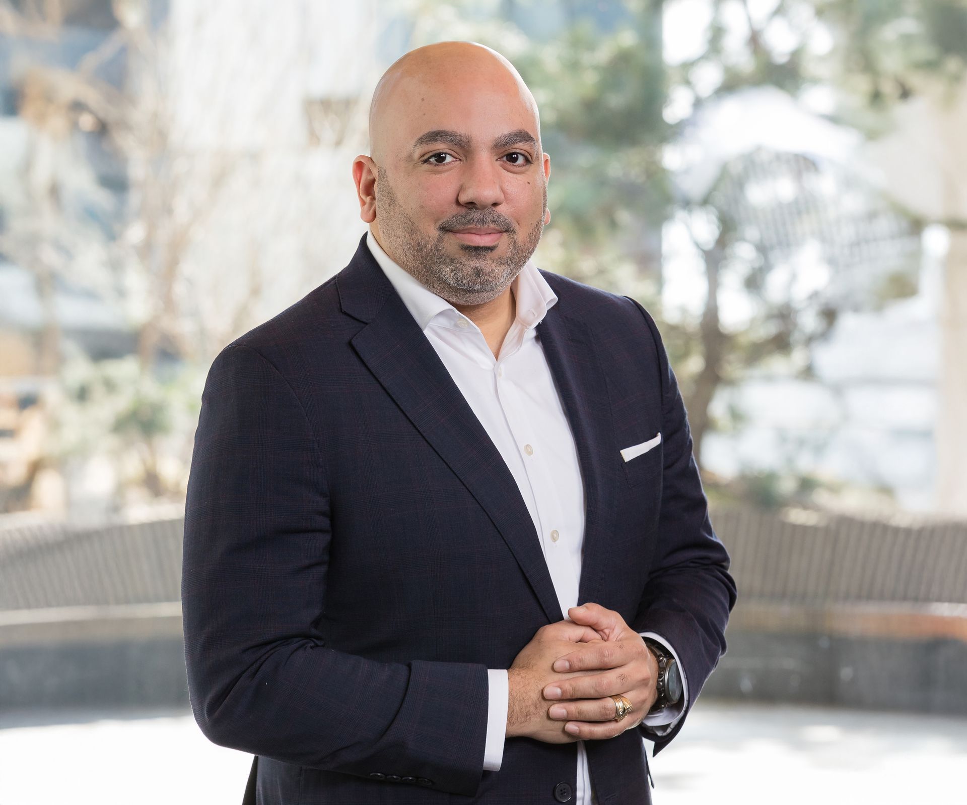 A bald man in a suit and white shirt is standing with his hands folded.