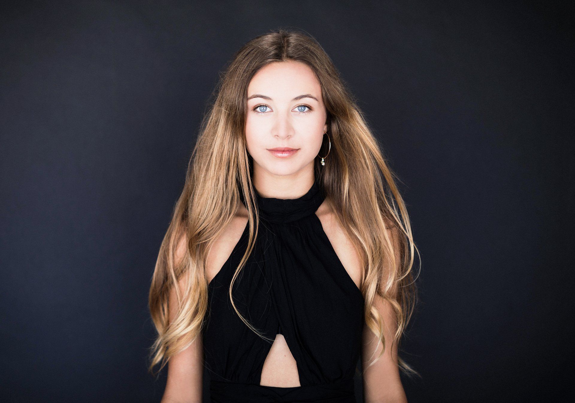 A young woman in a black dress is standing in front of a black background.