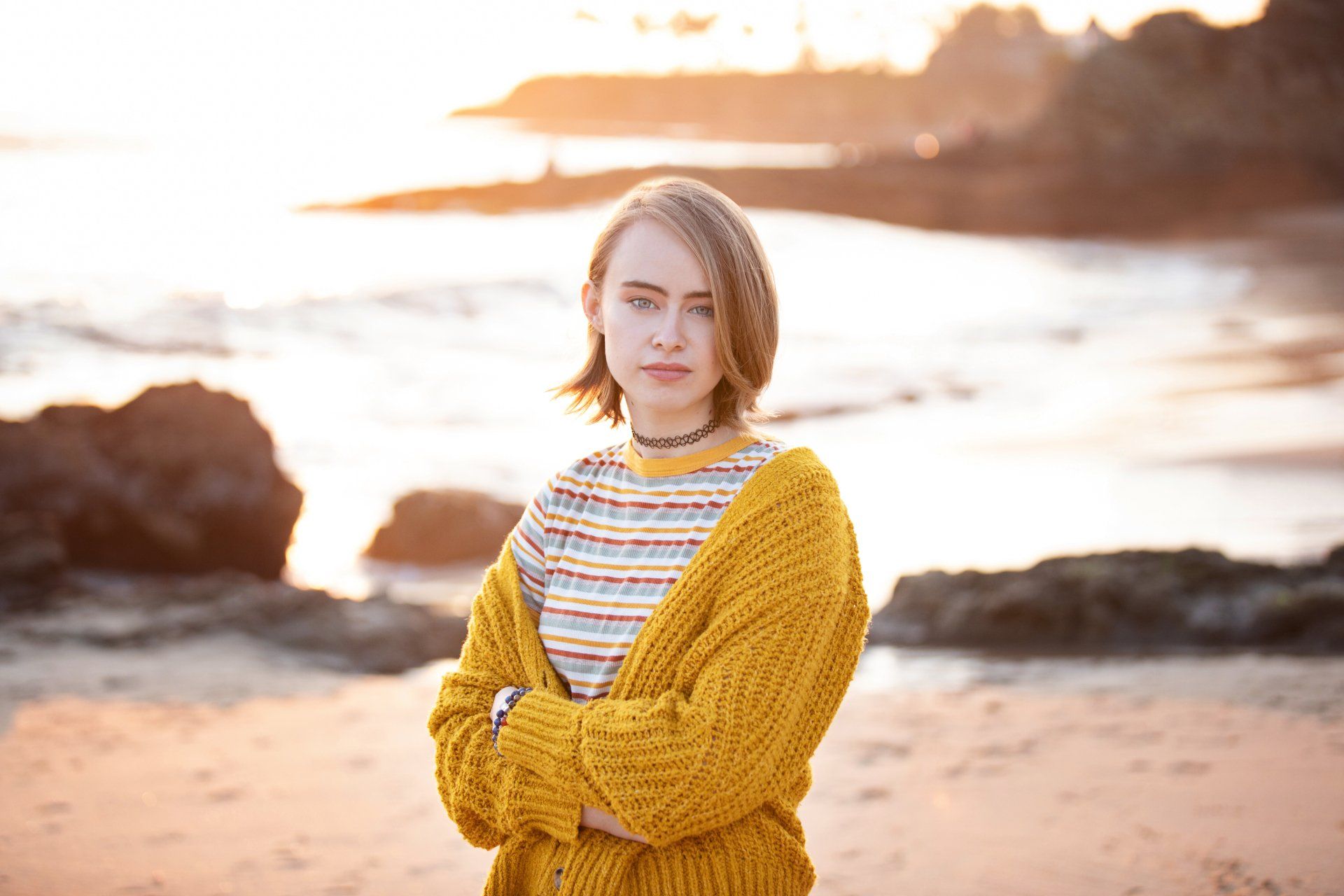 A woman in a yellow sweater is standing on a beach with her arms crossed.