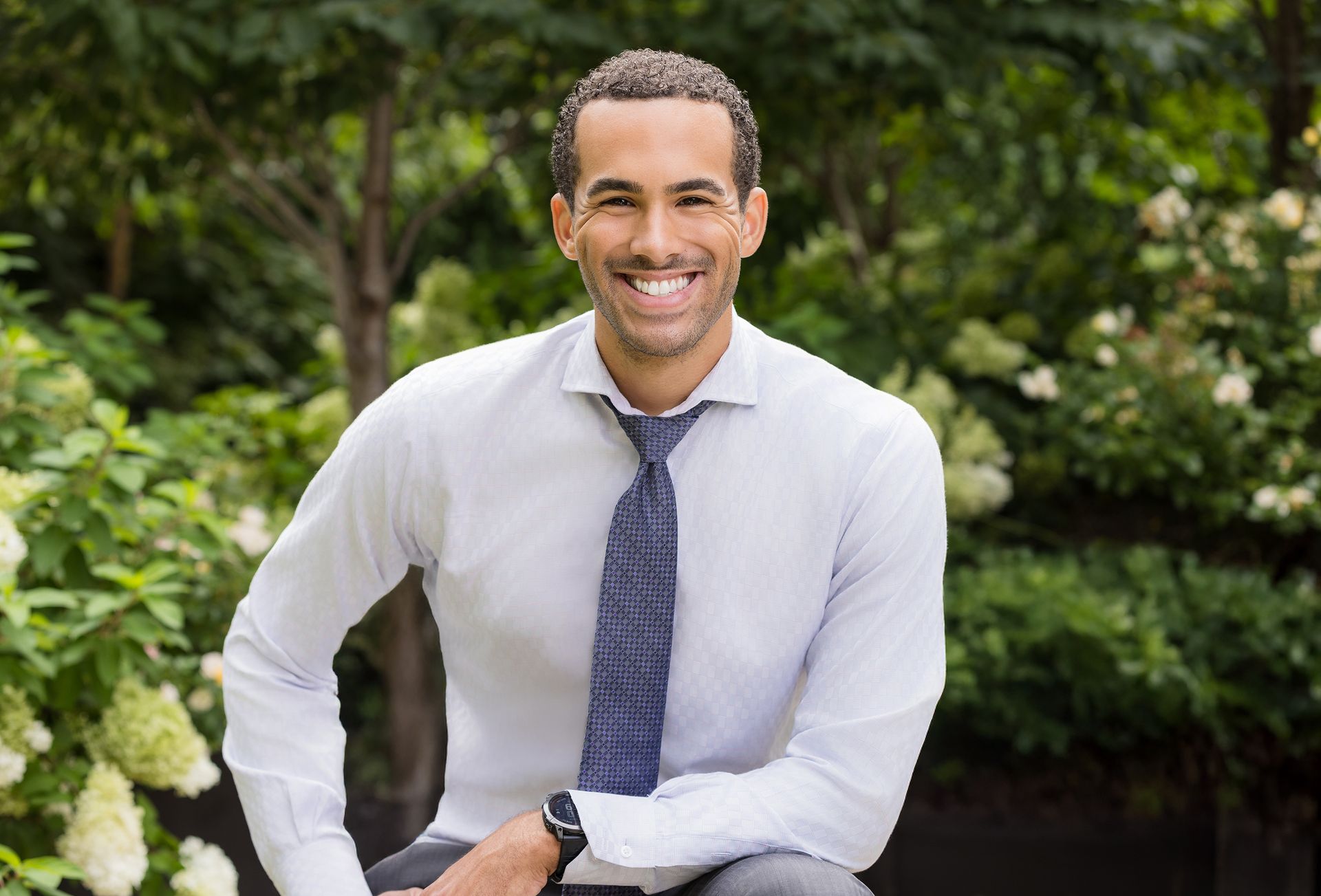 A man in a white shirt and tie is sitting in front of a bush.